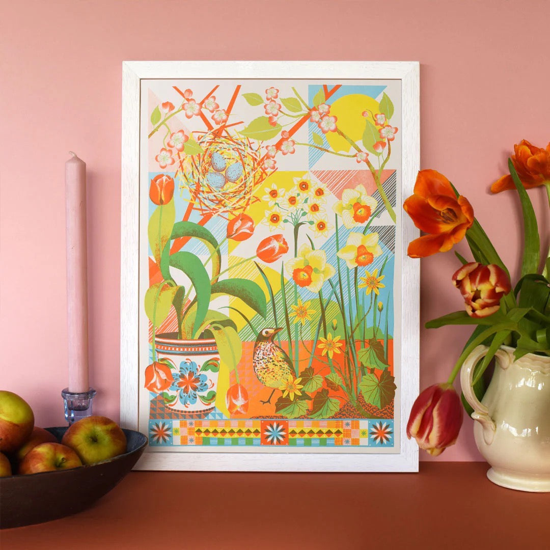Colorful floral and bird illustration in a white frame on a pink wall with a bowl of apples and a vase of flowers.
