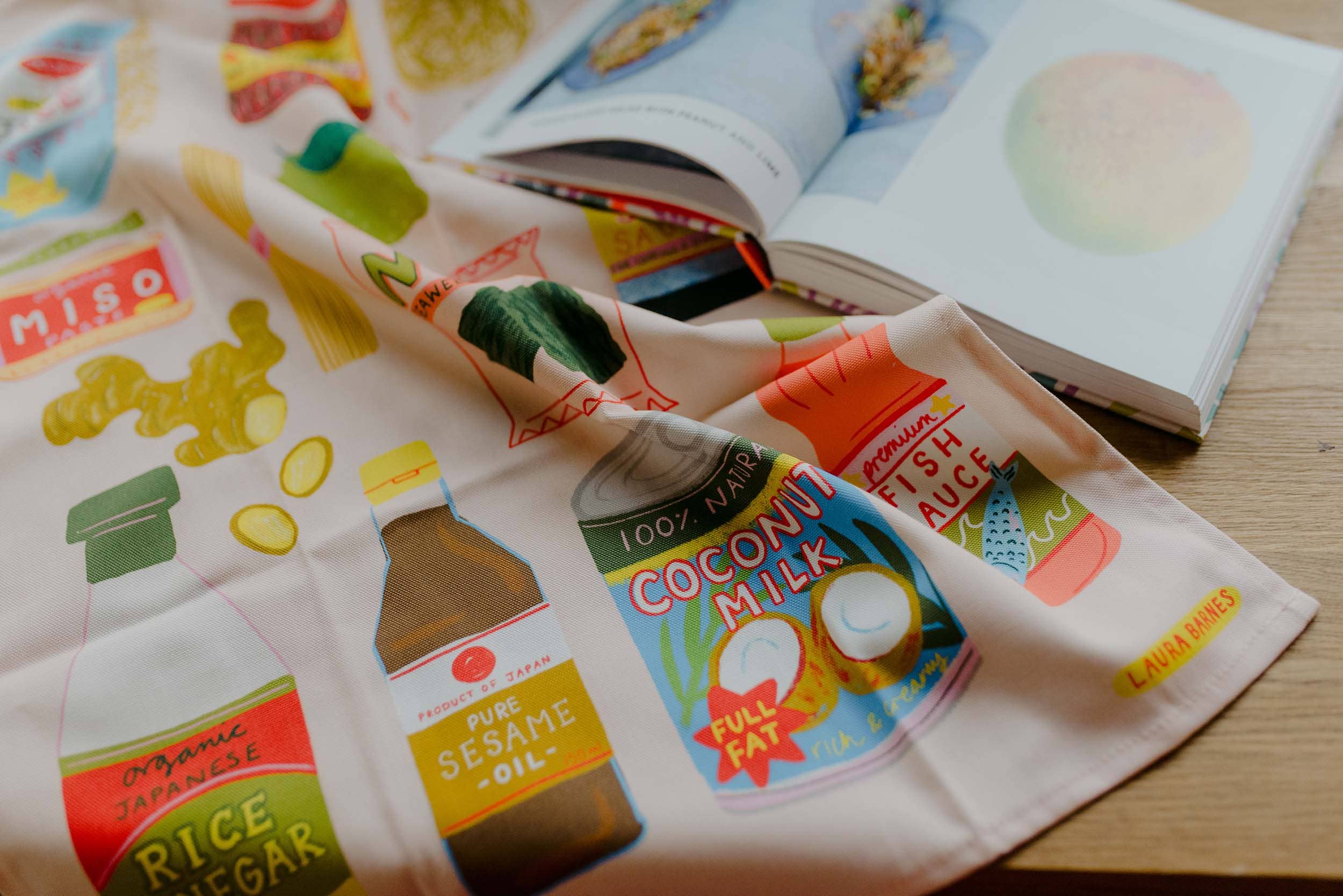 Tea towel with food-themed designs including coconut milk, rice vinegar, and sesame oil on a wooden surface.