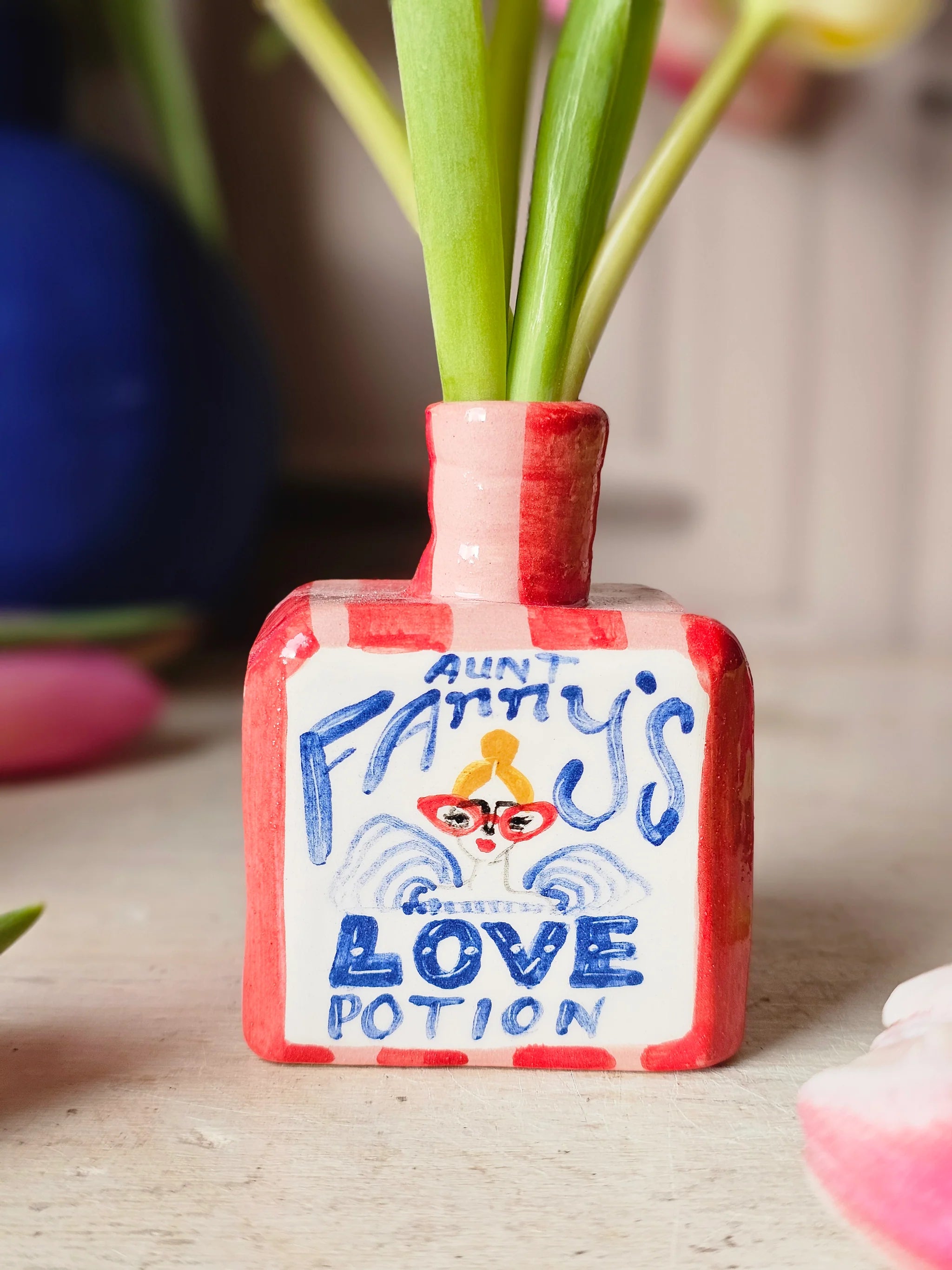 Small red and white vase with 'Fanny's Love Potion' text and flowers on a wooden surface.