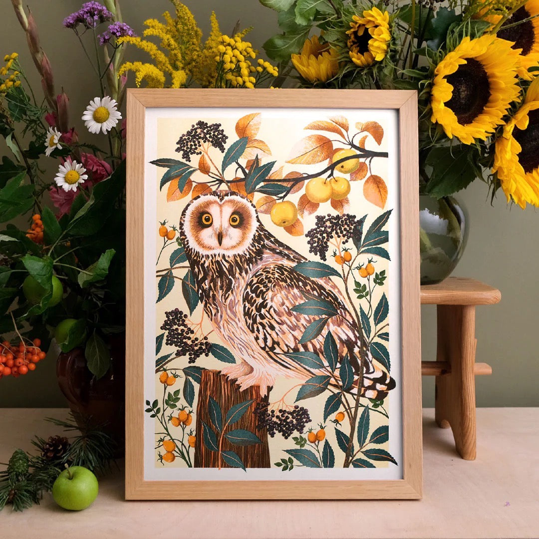 Framed artwork of an owl surrounded by flowers and fruits on a wooden surface.