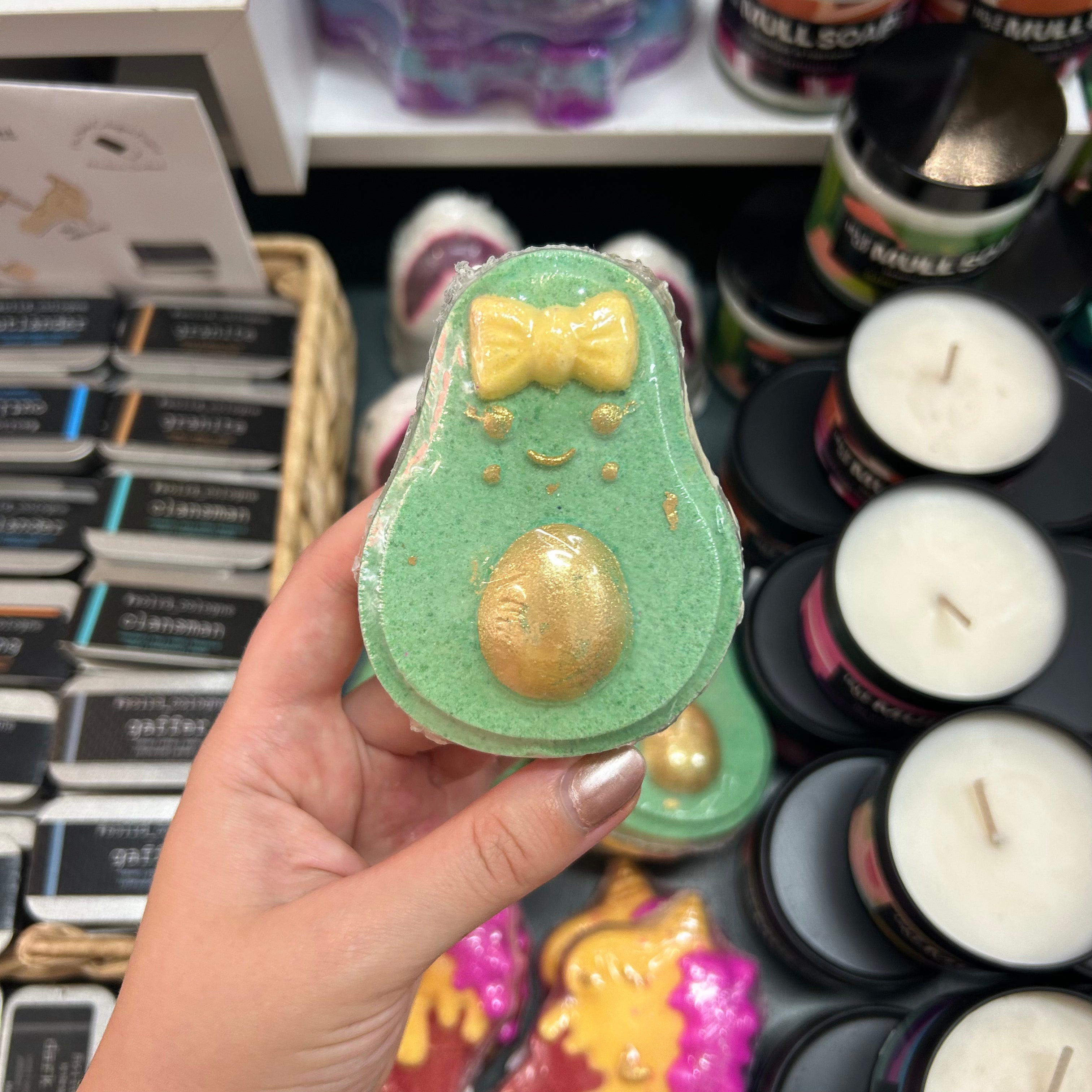 A hand holding a green avocado-shaped bath bomb with a yellow bow-tie and gold glitter belly, with candles and packaged soaps visible in the background.