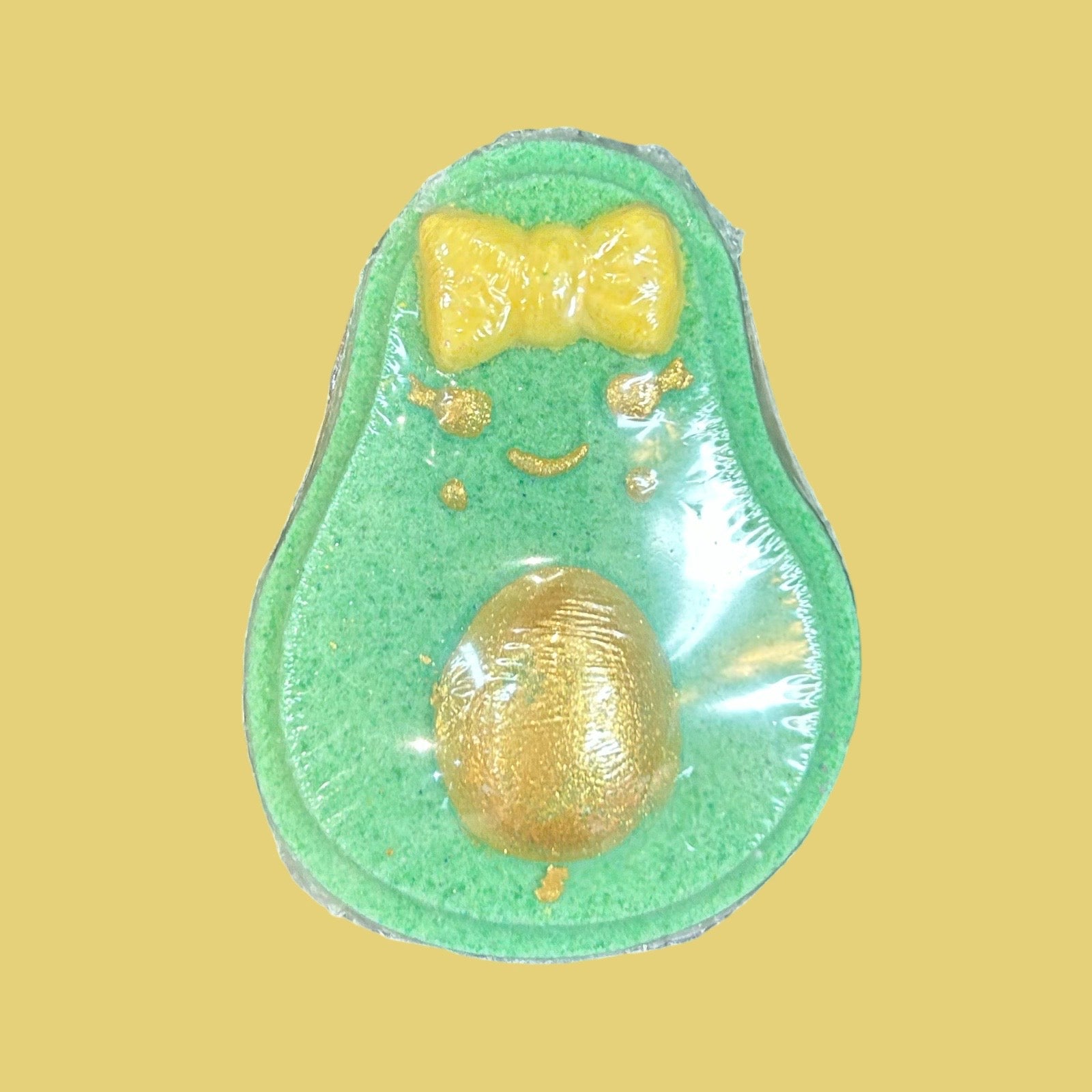 A green avocado-shaped item with a shiny gold glitter pit and a yellow bow-tie detail, centered on a solid pale yellow background.