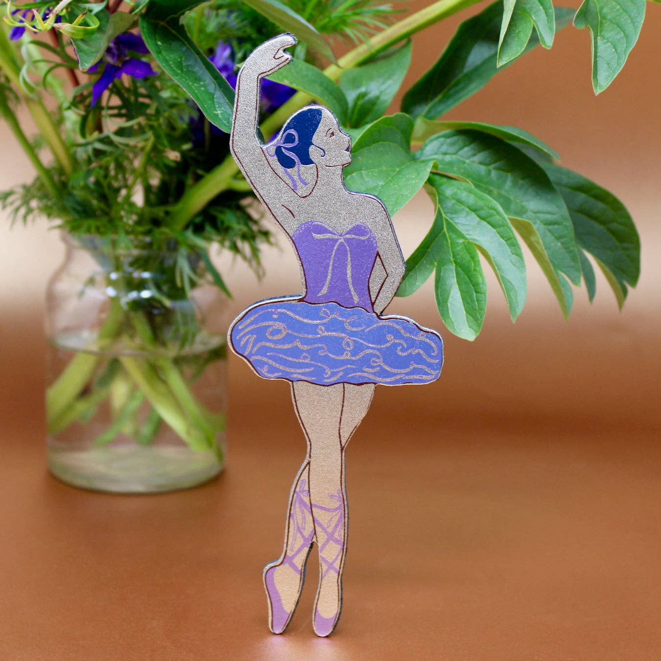 Decorative ballerina figure bookmark with a purple tutu on a wooden surface with green leaves in the background.