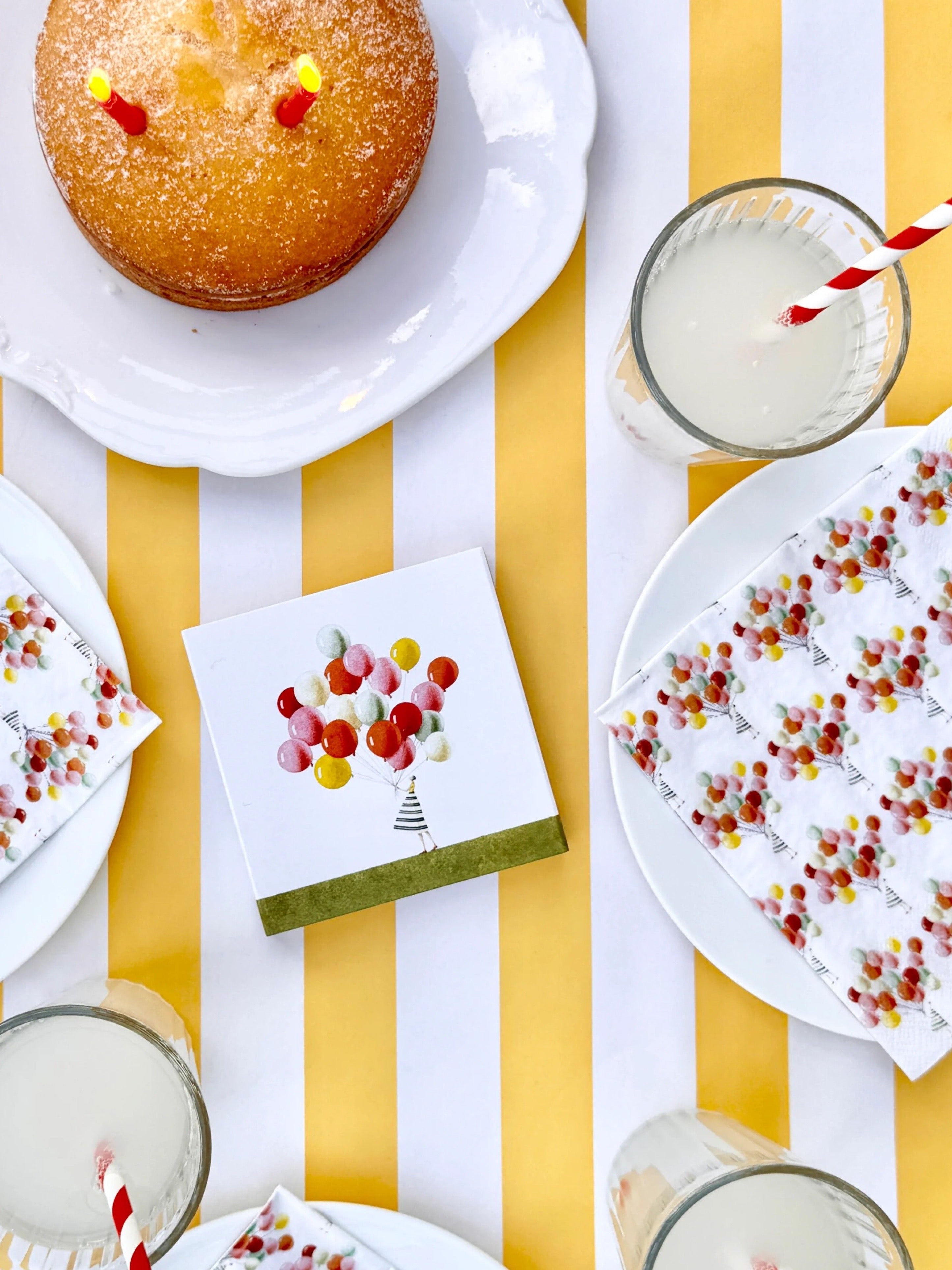 Table setting with a cake, drinks, napkins, and a card on a yellow and white striped tablecloth.