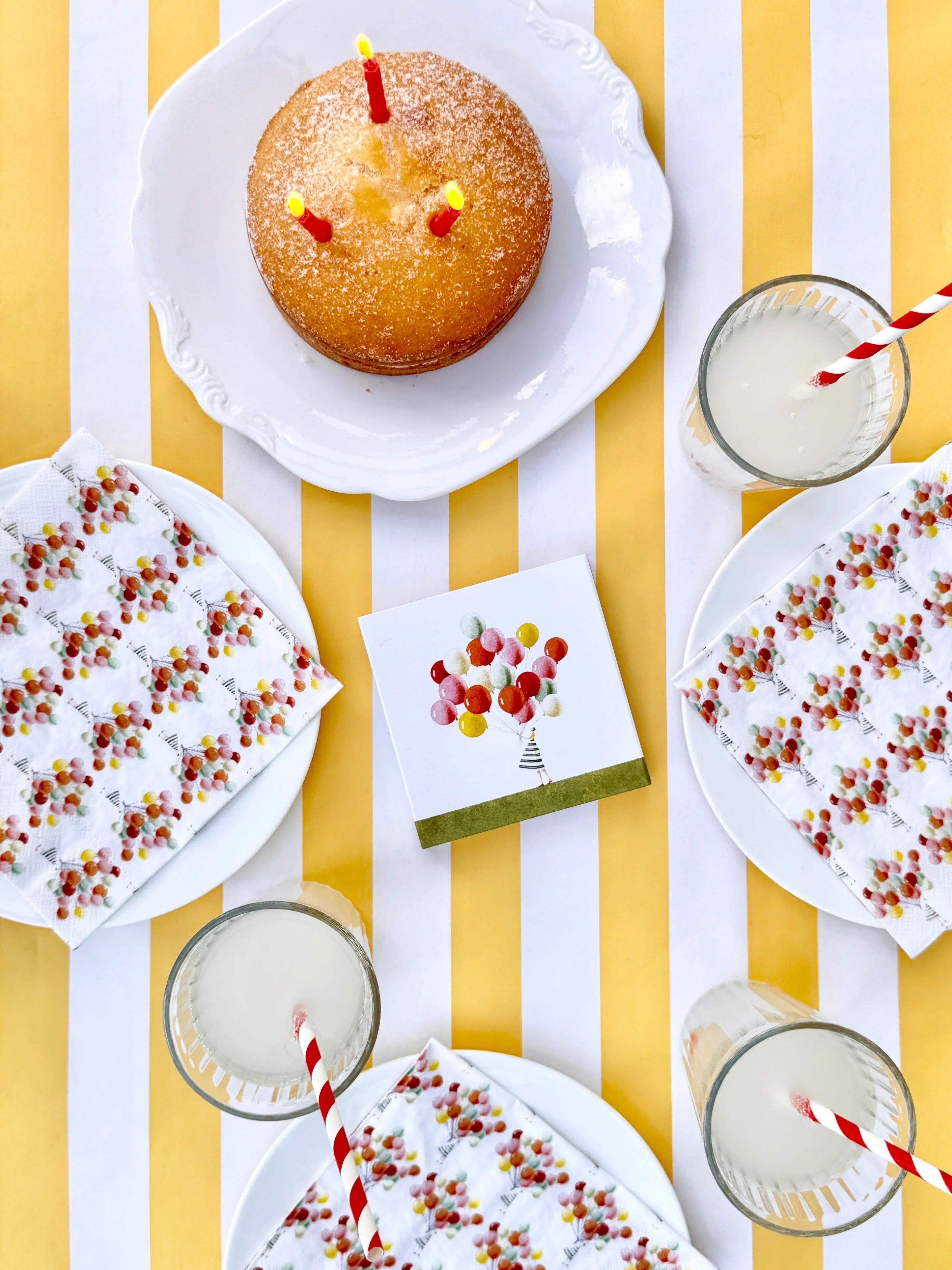 Cake with candles on a striped yellow and white background with napkins and drinks.