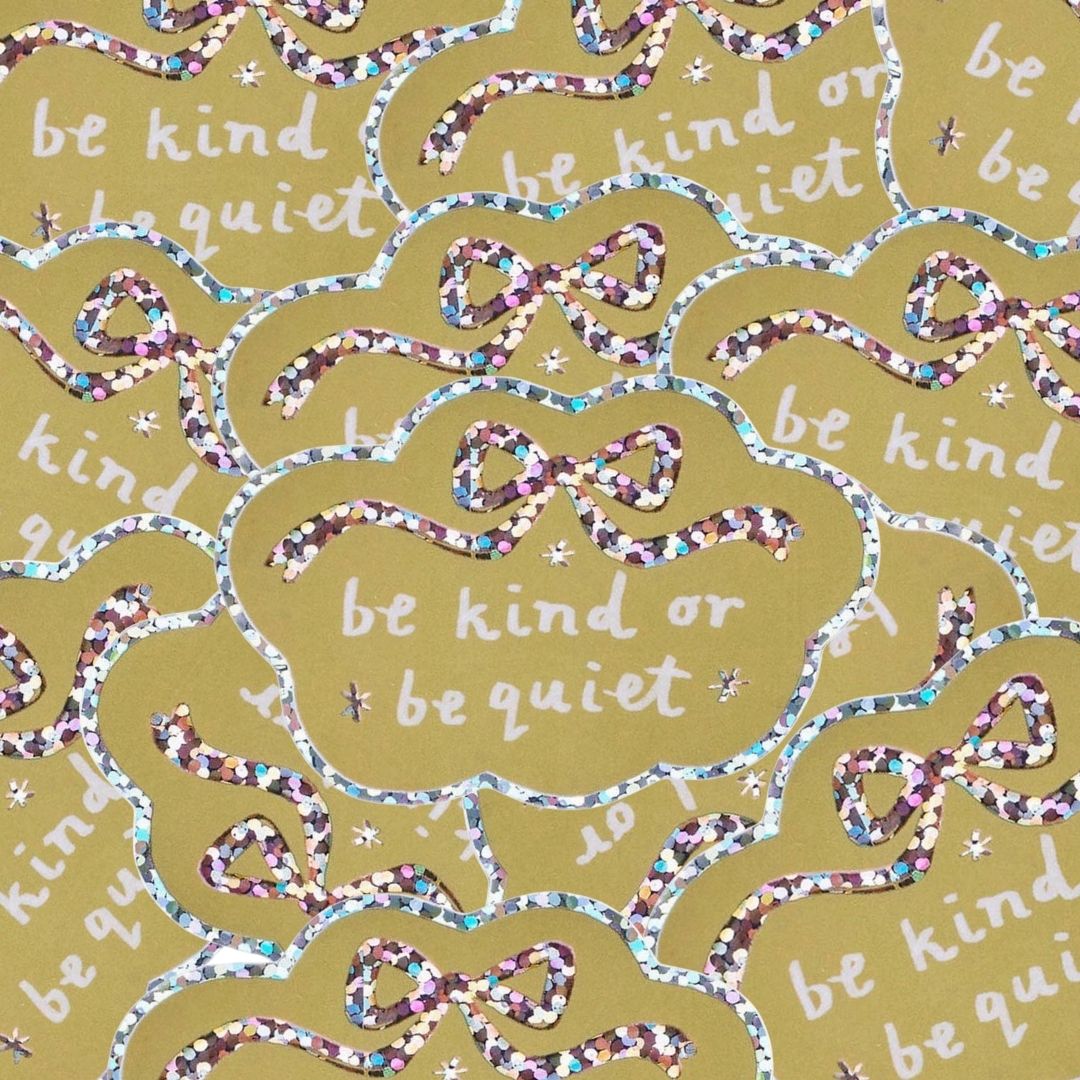 A close-up of a pile of stickers in yellow cloud-like shapes with the phrase 'be kind or be quiet' in white script, outlined with glittery ribbon bows in multicolour.