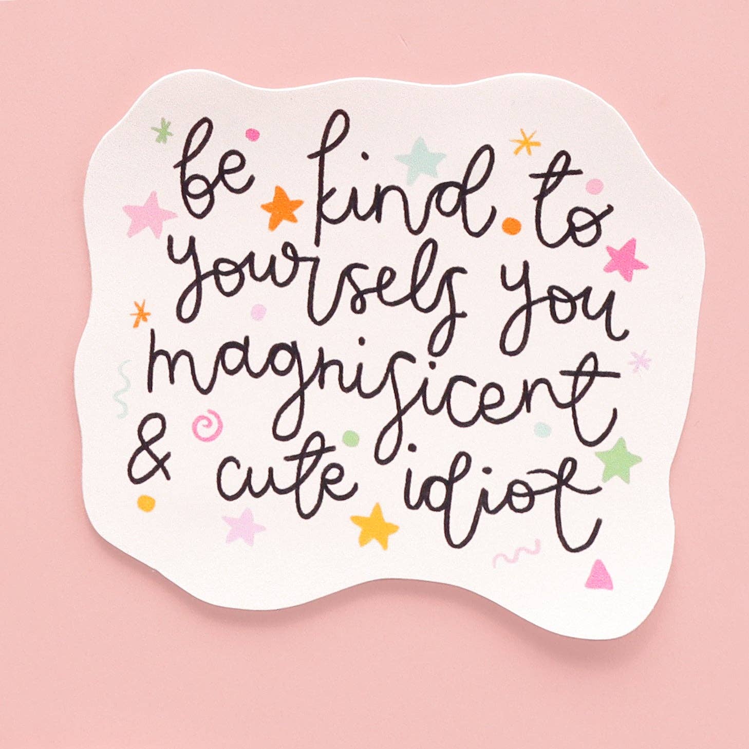 Motivational sticker with text 'be kind to yourselves you magnificent & cute idiot' on a pink background.