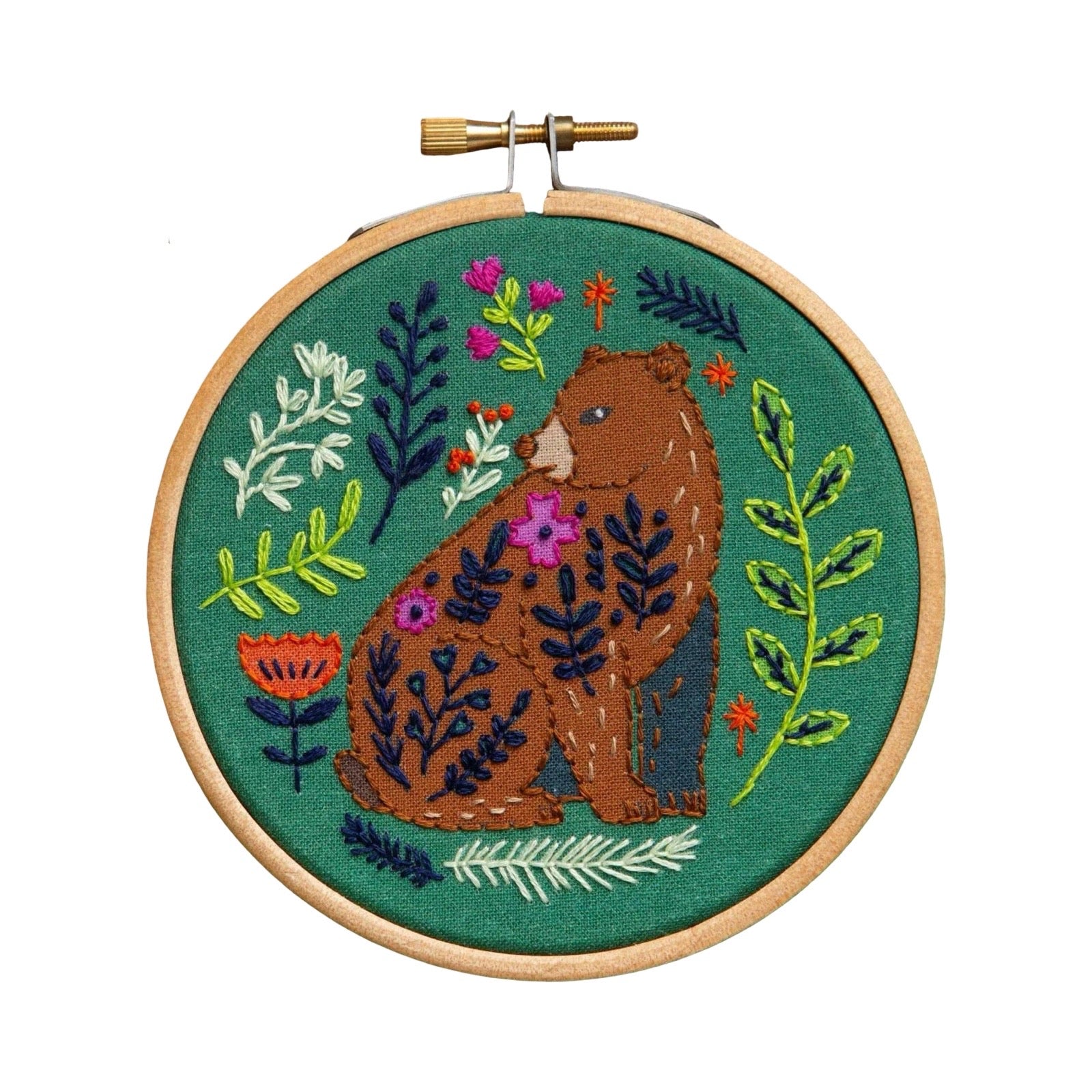 A circular embroidery hoop containing a finished embroidered design of a brown bear surrounded by green leaves and small colorful flowers on a teal-green background, photographed against a plain white background.