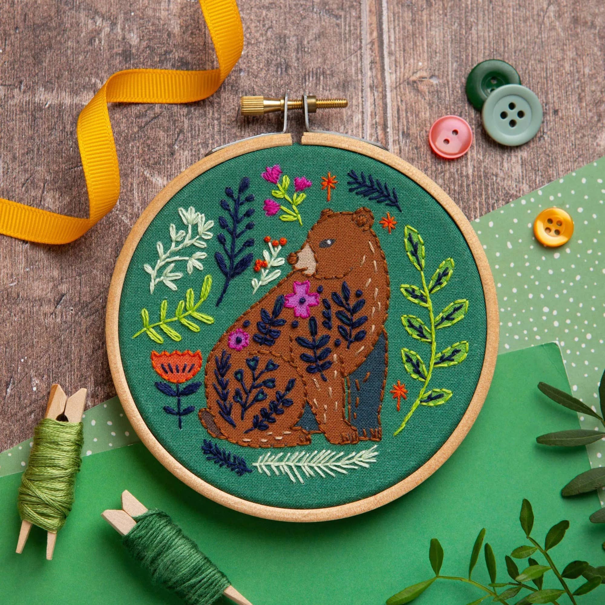 A round wooden embroidery hoop displaying a finished embroidered design of a brown bear surrounded by colourful flowers and leaves on a green fabric background. The hoop is styled on a rustic wood surface with a yellow ribbon, green thread spools, decorative buttons, and green paper.