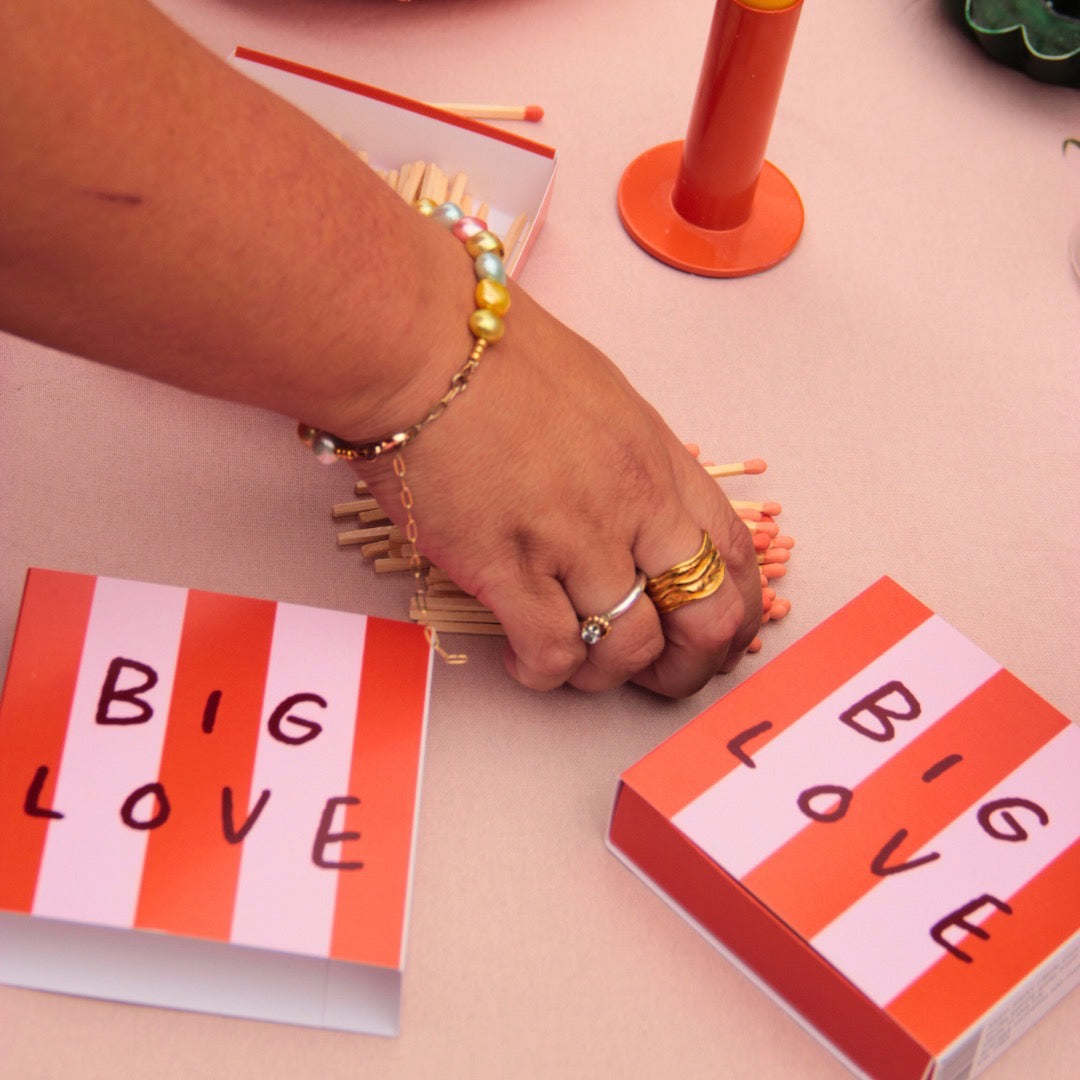 This matchbox features a red and pink striped design with the text 'Big Love', on a table. 