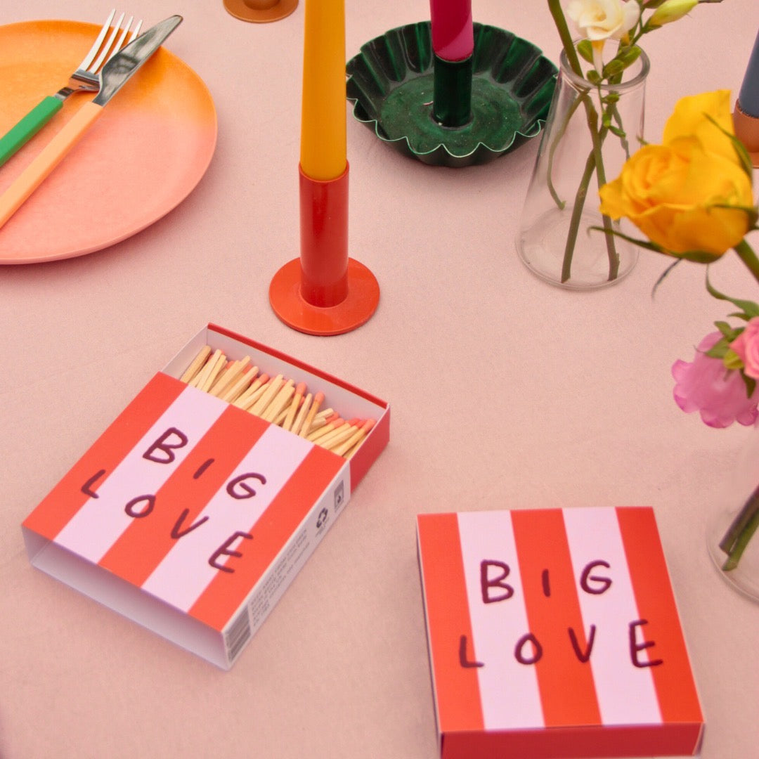 This matchbox features a red and pink striped design with the text 'Big Love', on a table.