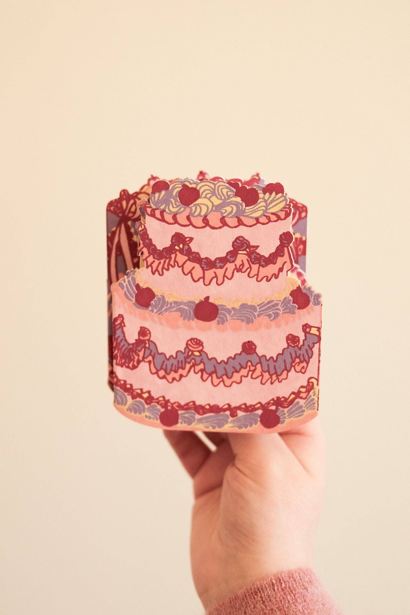 Hand holding a small, decorative cake-shaped card with intricate designs on a beige background.