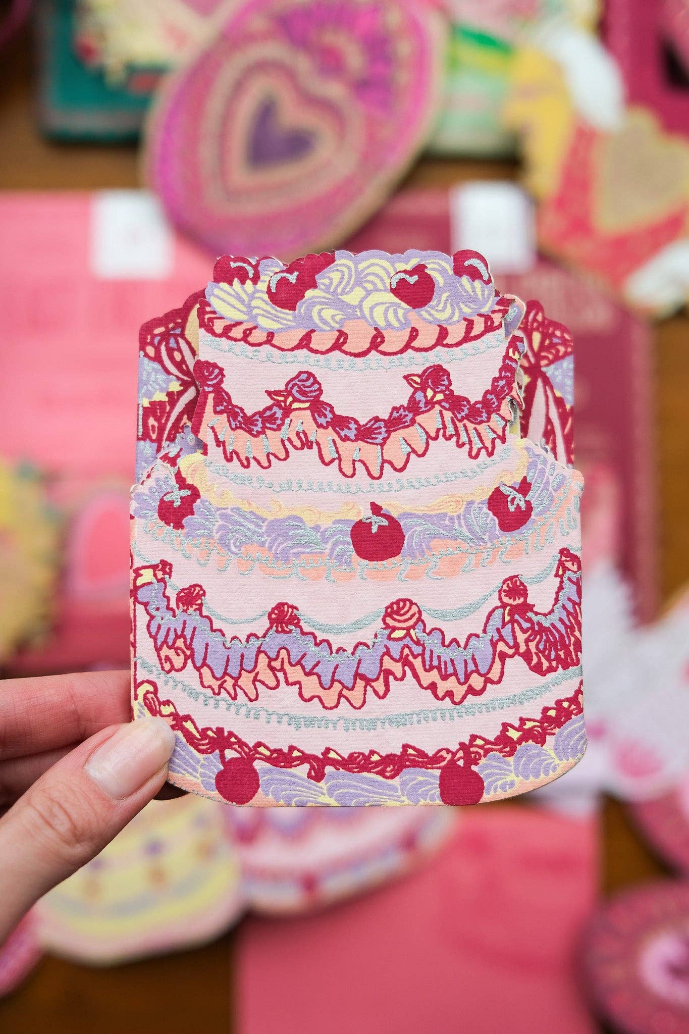 Colourful birthday cake card with decorative elements held by a hand against a blurred background.