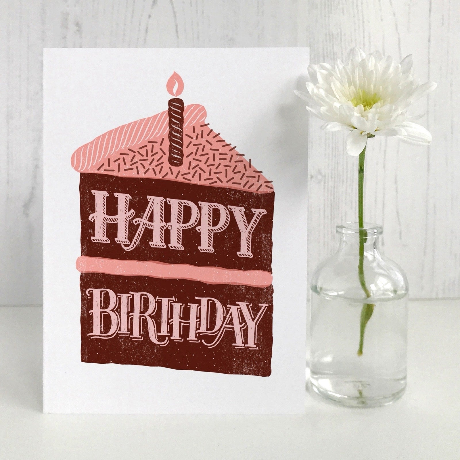 Birthday card with cake illustration and 'Happy Birthday' text next to a vase with a flower on a light wooden surface.