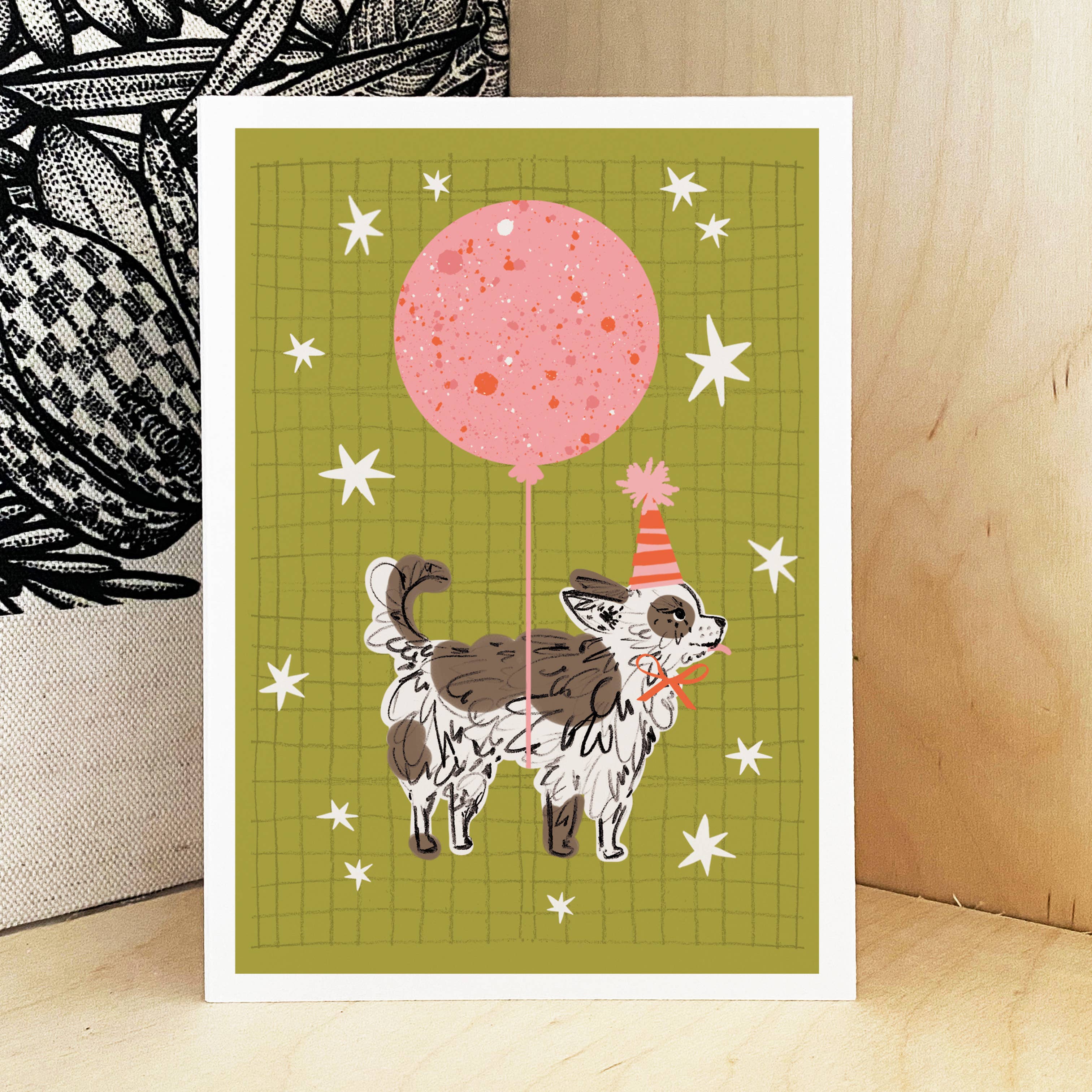 A greeting card with a white border, featuring a whimsical illustration of a small white and brown dog wearing an orange party hat and holding a pink balloon, set against an olive green grid background with white stars. The card is displayed standing upright on a light wood surface next to a patterned textile.