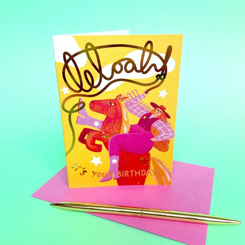 Colourful birthday card with cartoon characters on a pink envelope and gold pen against a teal background.