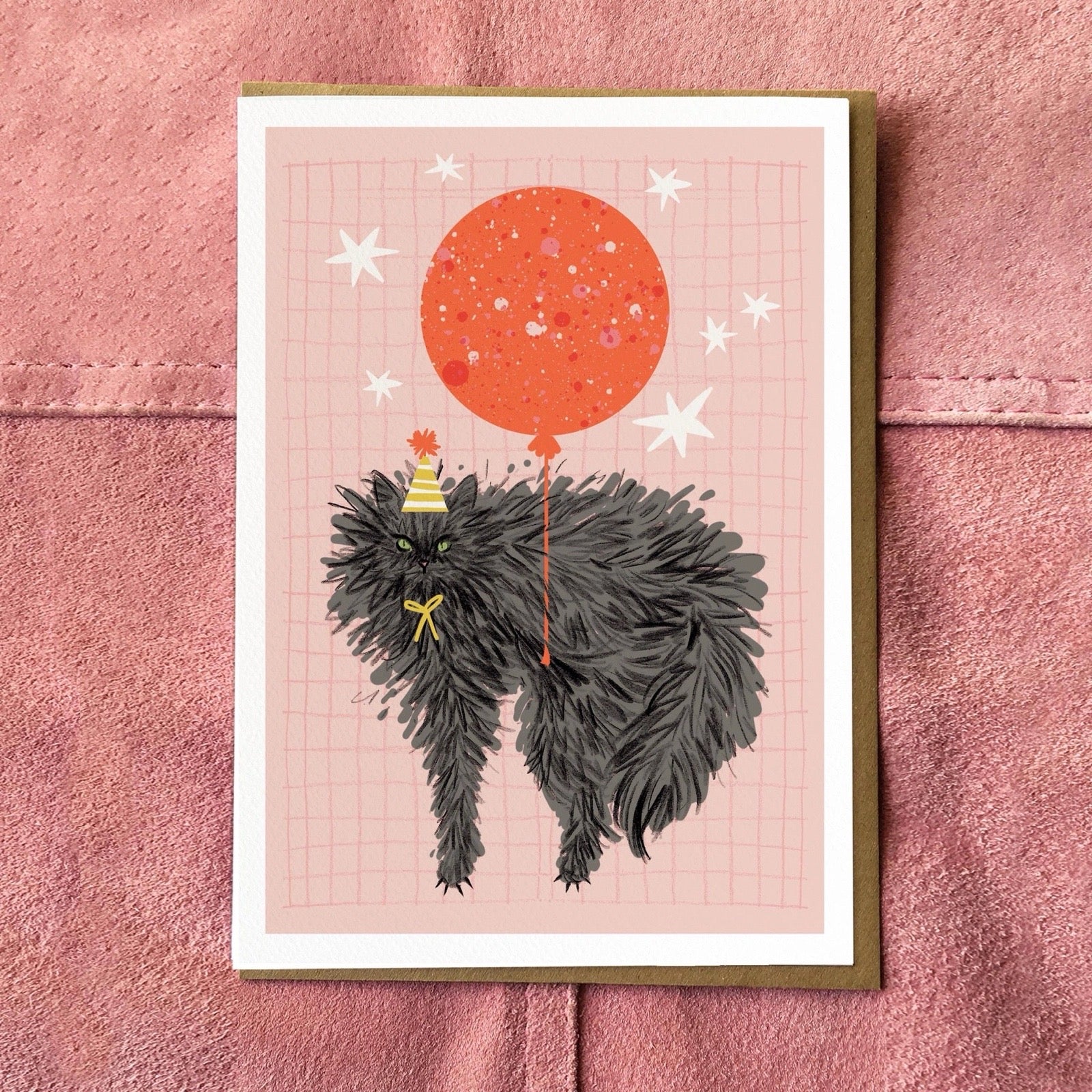 Illustration of a cat with a red balloon on a card, sat on a pink fabric background.