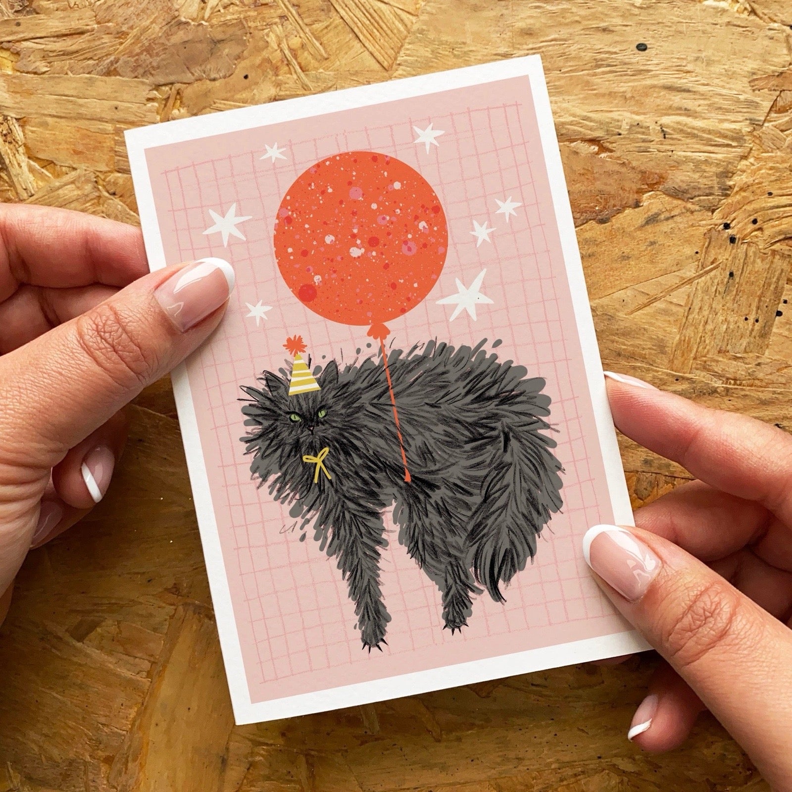 Card with a black cat and red balloon illustration held by hands on a wooden surface.