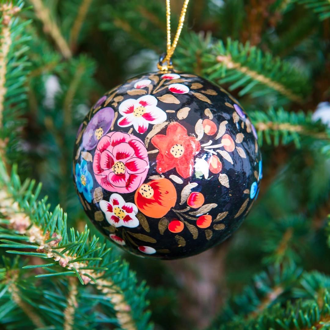 A round papier mache bauble in black with a colourful floral design hanging on a Christmas tree.