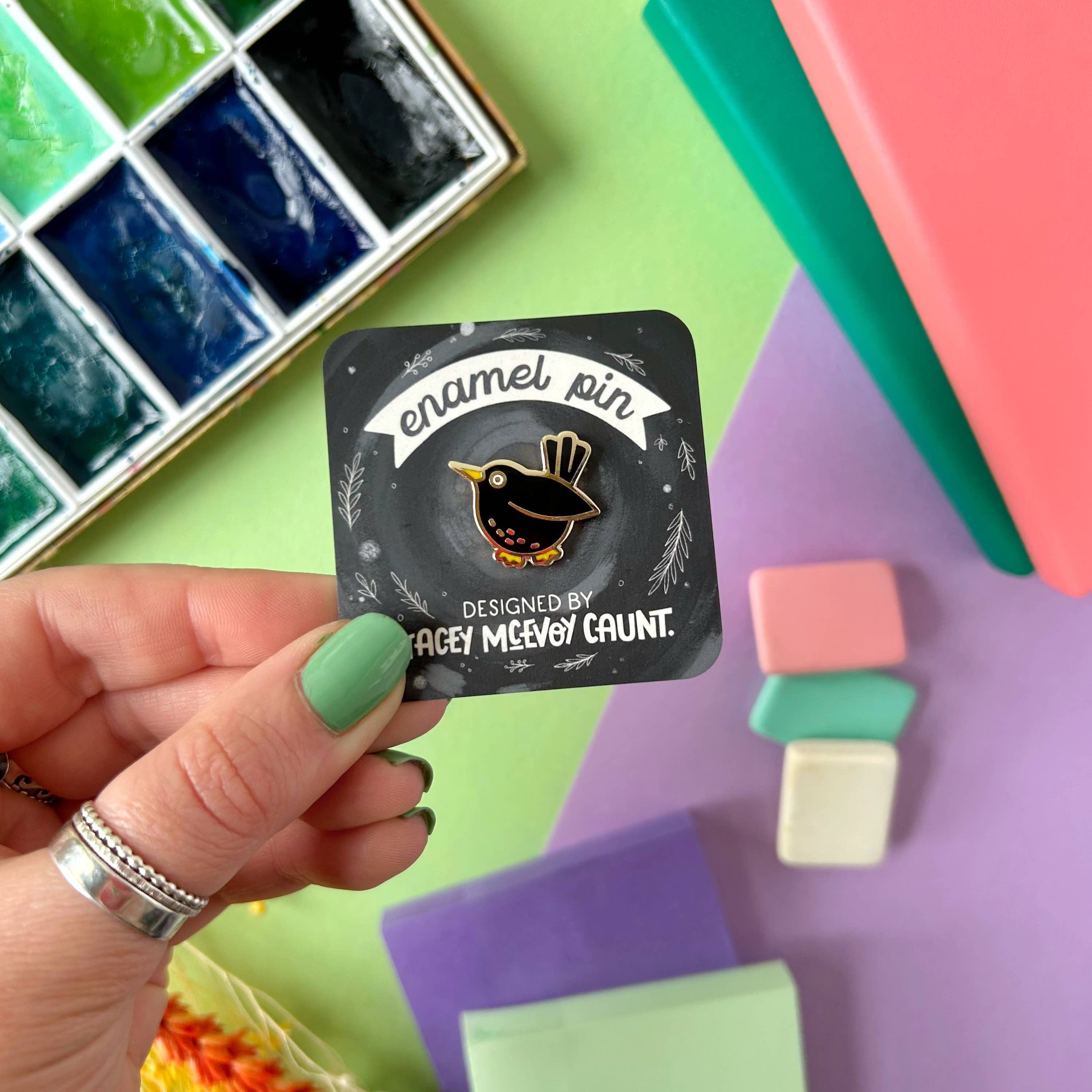 Hand holding a card with an enamel pin design, surrounded by art supplies on a colourful background.