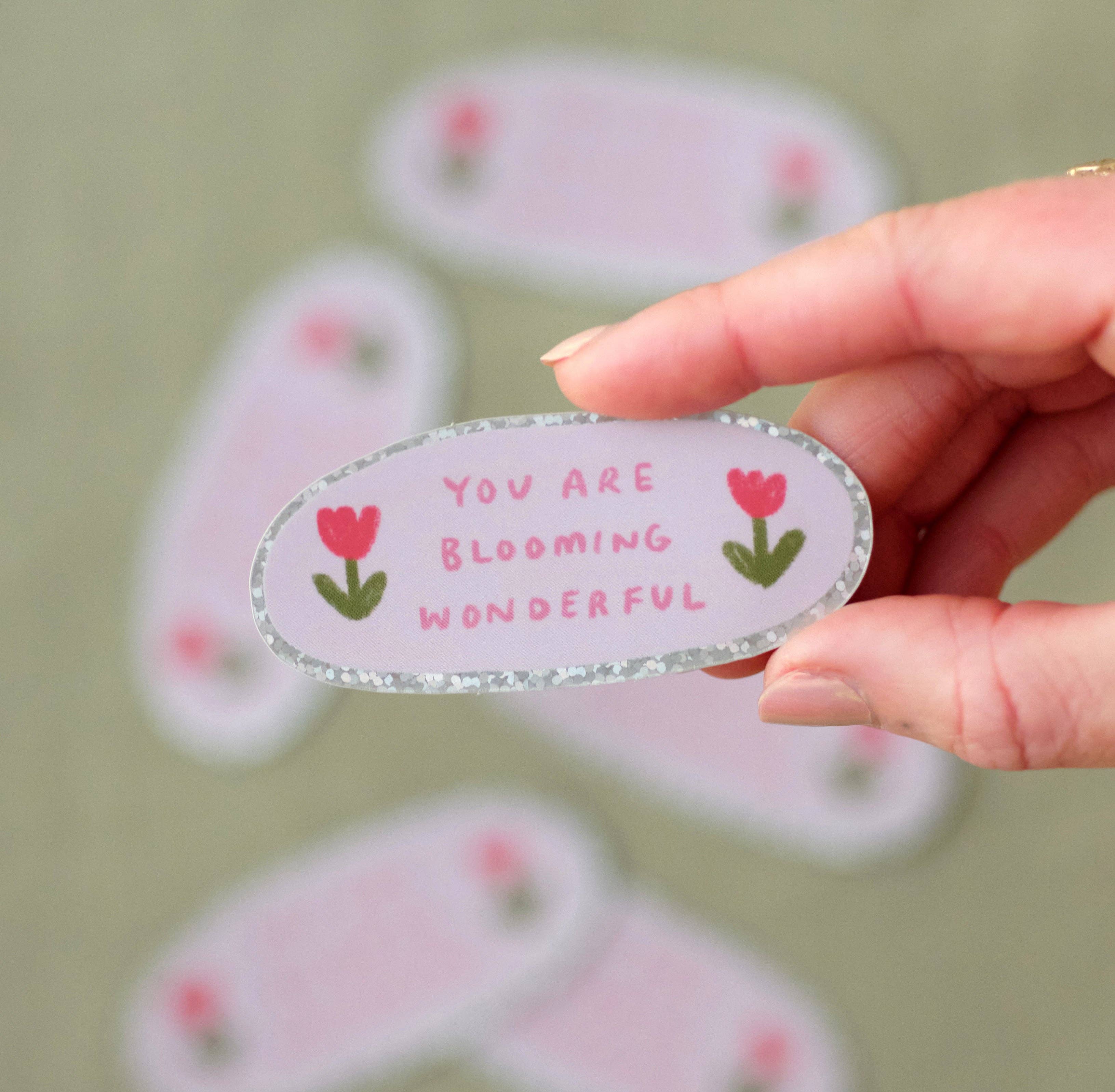 Hand holding a decorative sticker with 'You are blooming wonderful' text and flowers on a light green background.