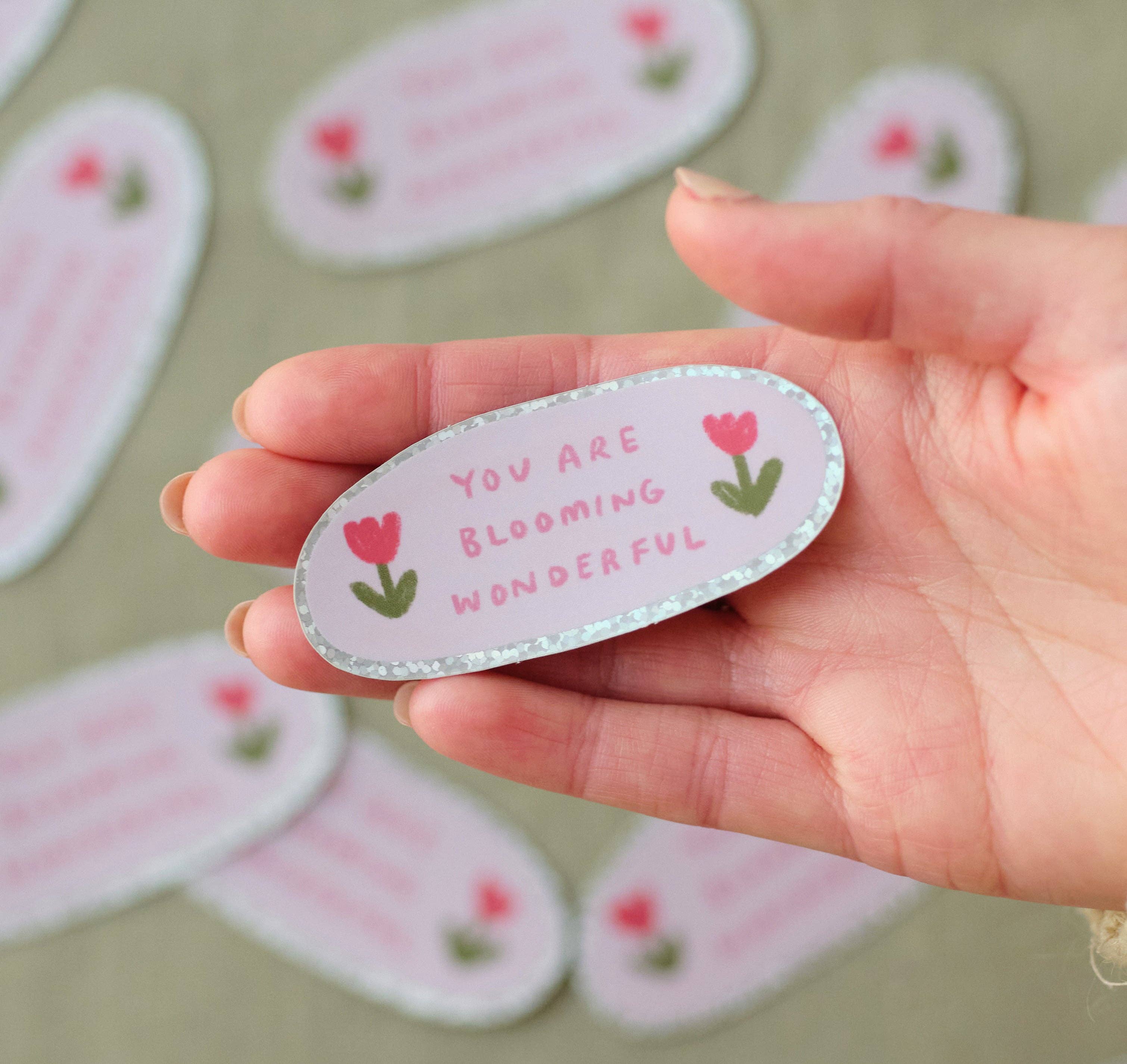 Hand holding a decorative sticker with 'You are blooming wonderful' text and flowers.