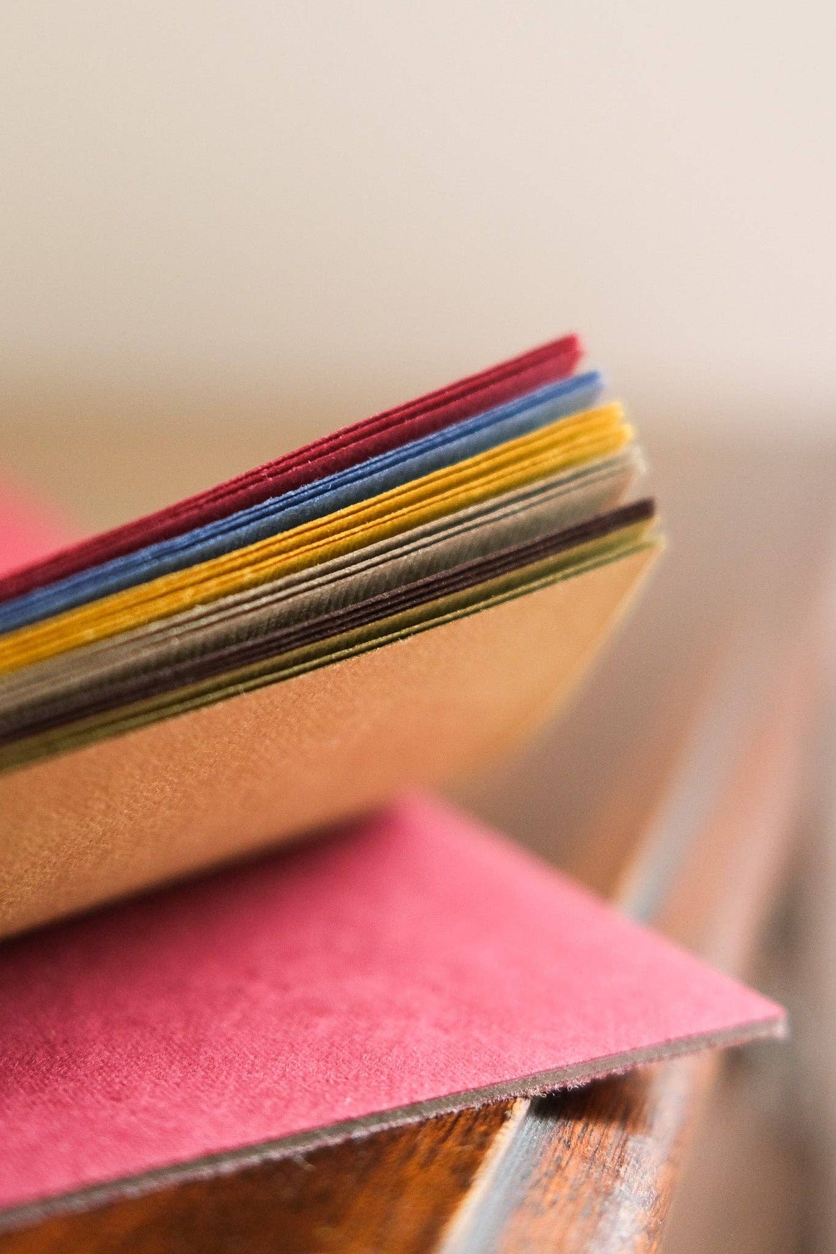 A close-up of a stack of colourful material or fabric swatches fanned out, with a pink sample resting on top, photographed on a wooden surface against a soft beige background.