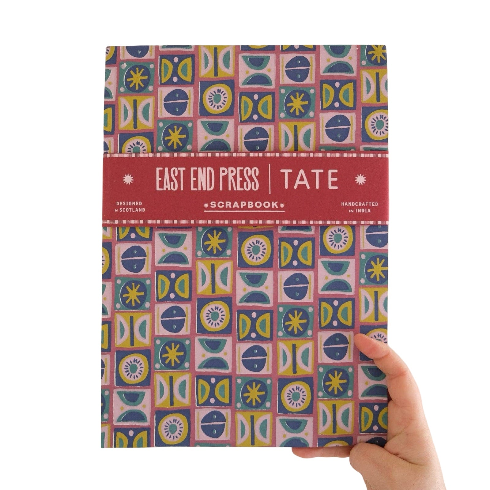 A hand holding a hardcover scrapbook book titled 'East End Press | Tate Scrapbook' against a plain white background. The book cover features a colourful retro geometric pattern with pink, blue, yellow, and green shapes, and a solid red horizontal band across the middle with white text.