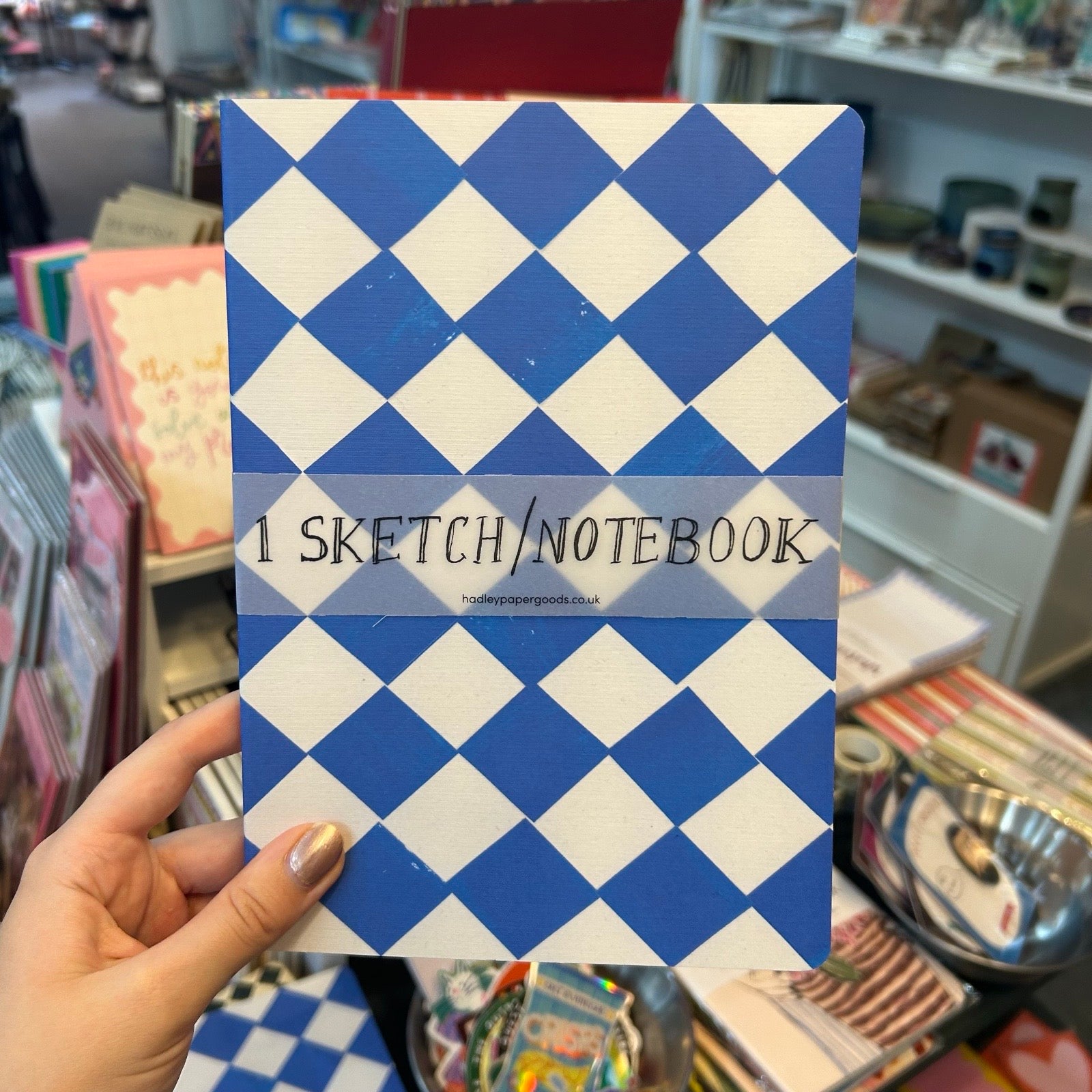 A hand holds a sketchbook with a blue and white diamond pattern cover in a shop setting. The cover has a semi-transparent banner across the middle that reads '1 SKETCH/NOTEBOOK' with a website address below it.