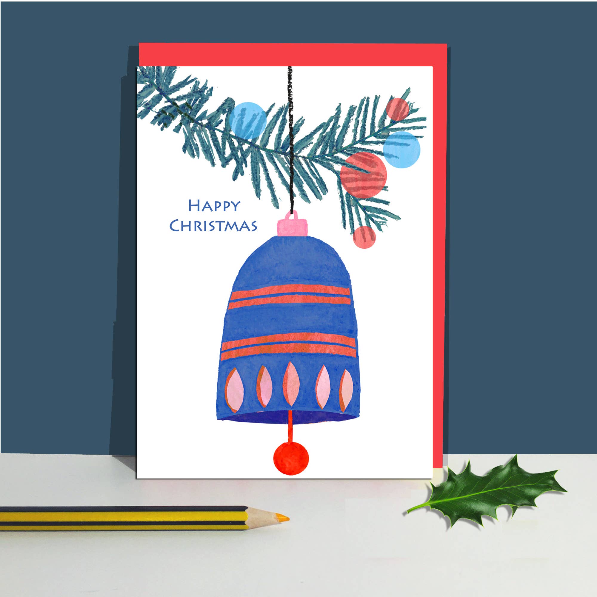 A Christmas greeting card with a white front and red envelope, featuring an illustration of a blue bell ornament with pink and red stripes, hanging pine branches, and the words 'Happy Christmas.' The card is photographed standing upright on a white surface against a dark blue background, with a pencil and a holly leaf.
