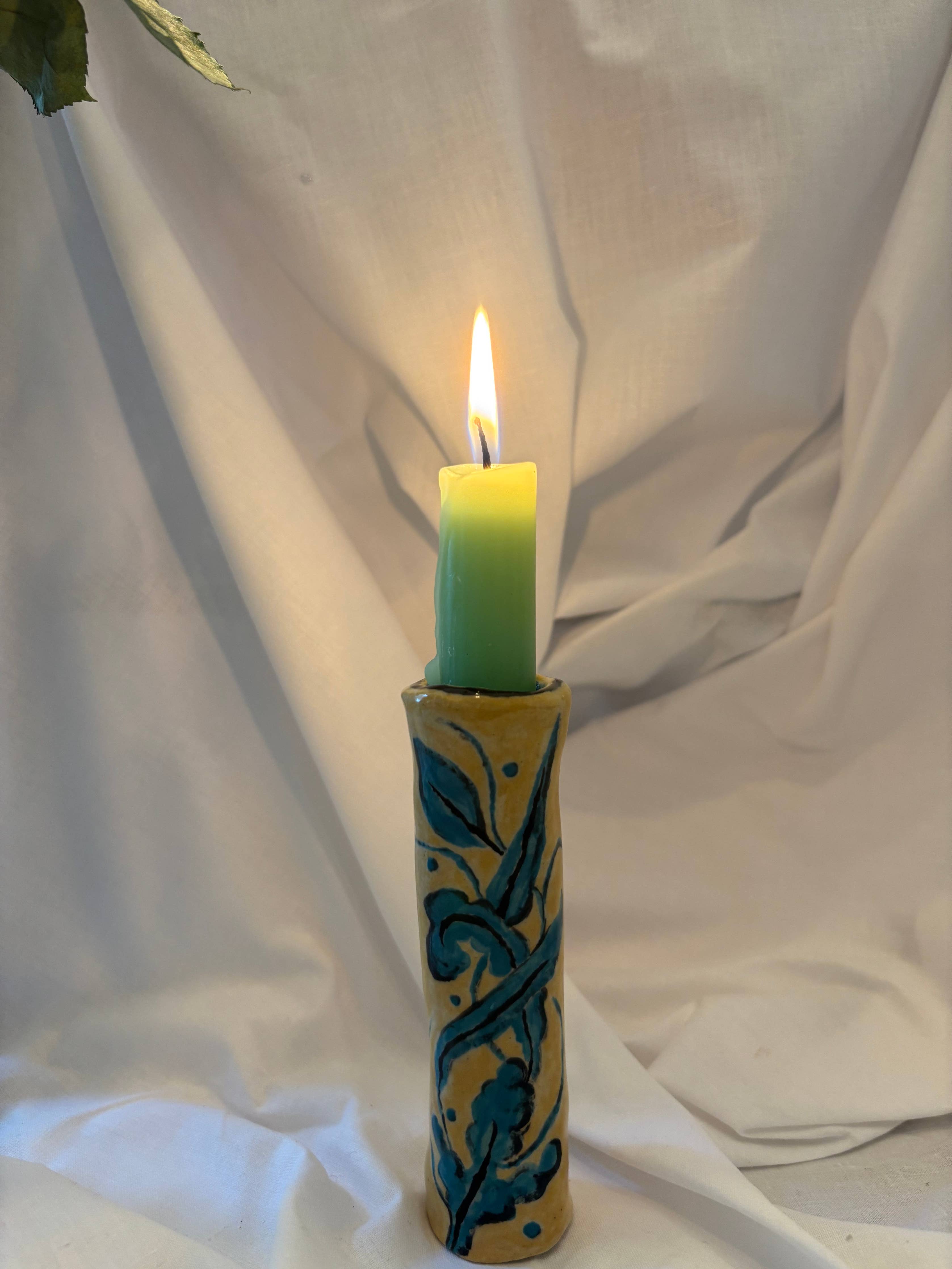 Candle in a decorative yellow holder with blue flowers on a white fabric background.