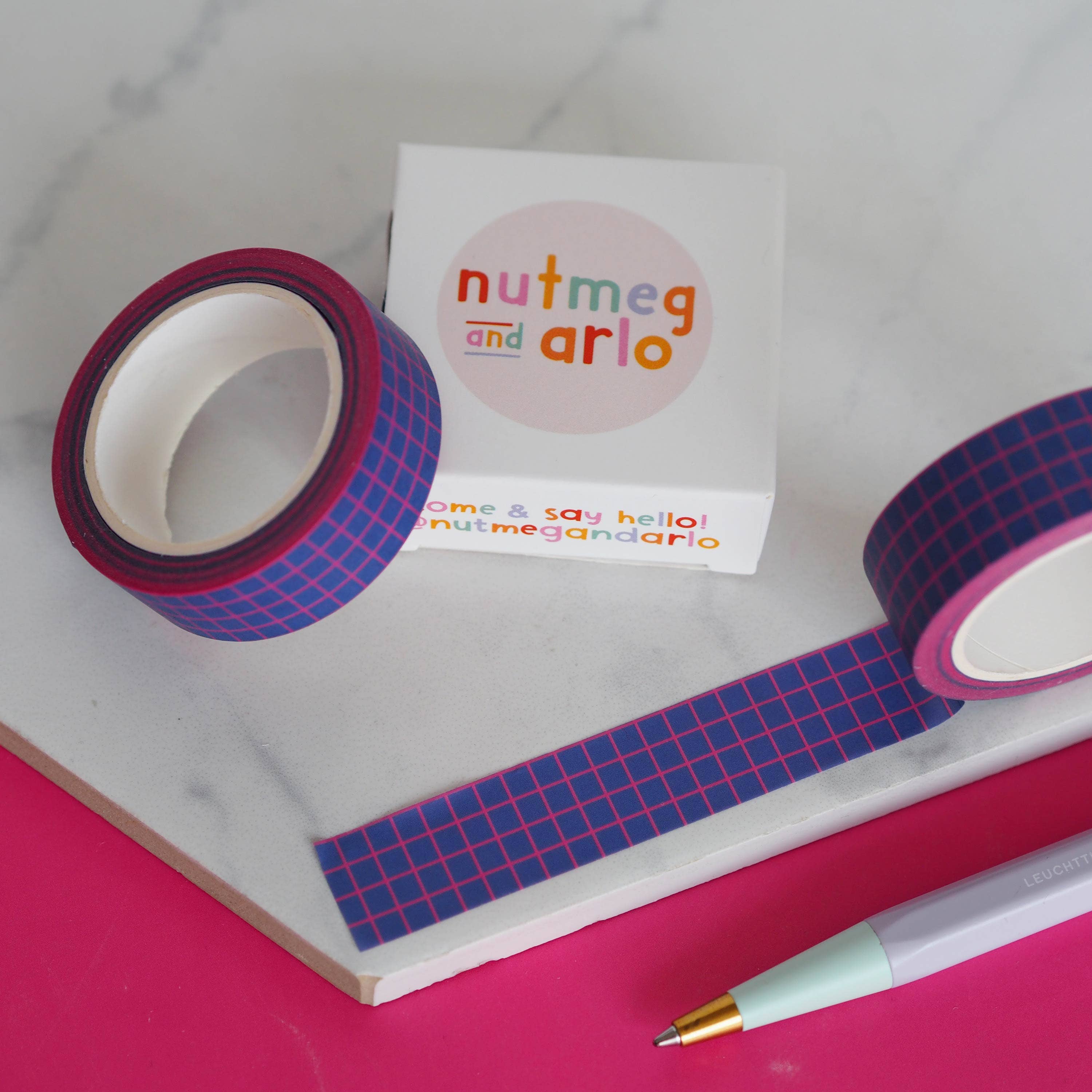 Rolls of colorful tape with a 'nutmeg and arlo' branded box on a pink surface.