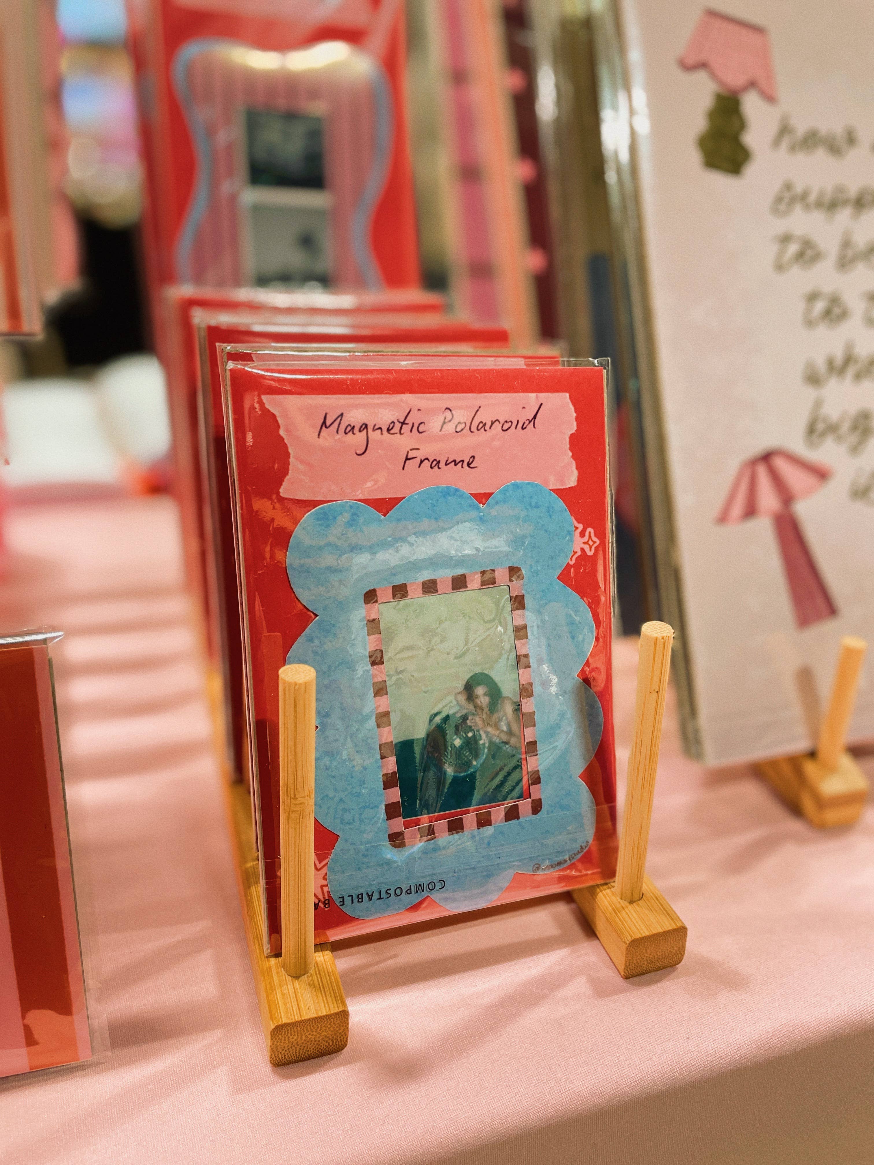 A stack of red and blue magnetic polaroid frames displayed on small wooden stands on a pink table, with a blurred store background.