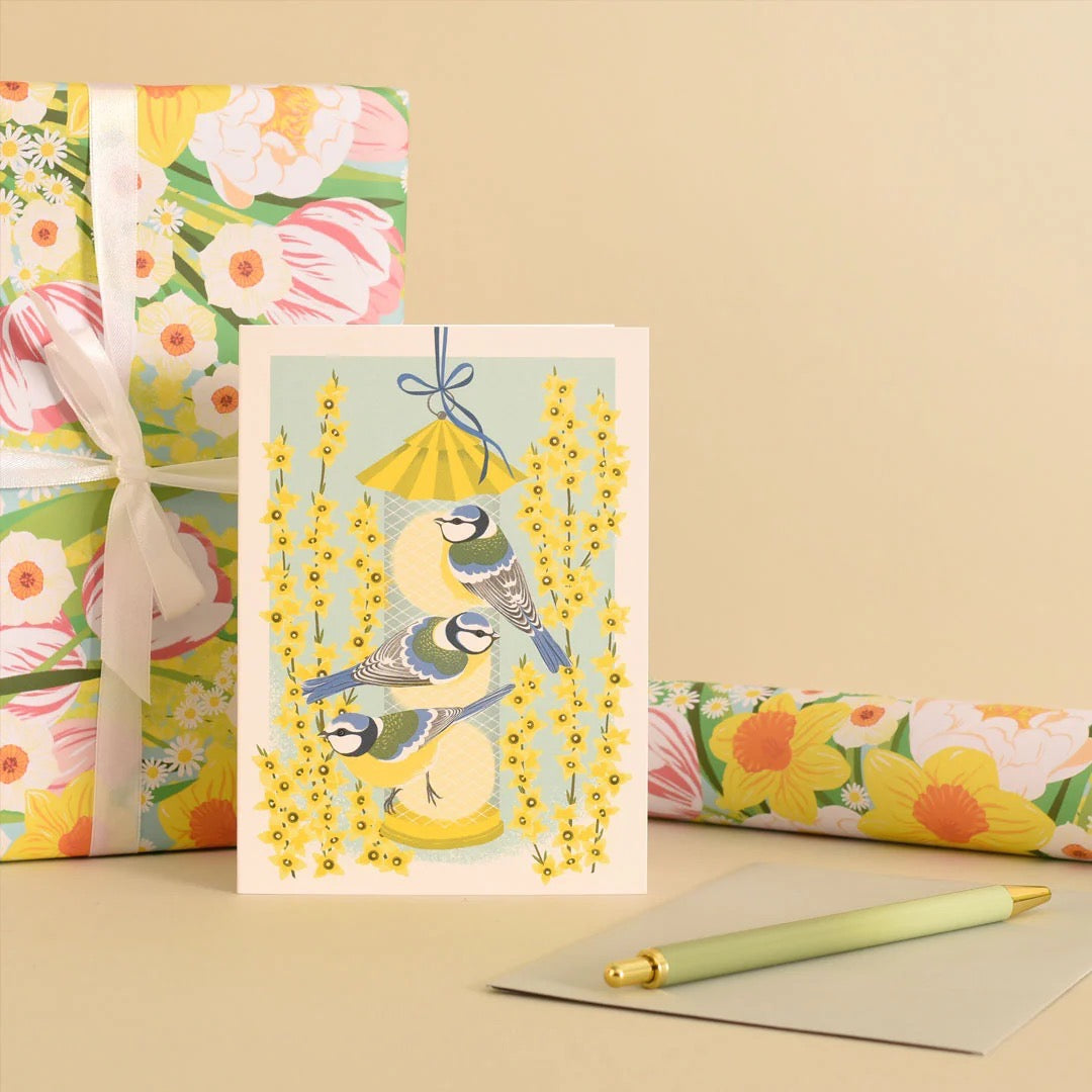Greeting card with birds and flowers on a beige background, surrounded by floral wrapping paper and a pen.