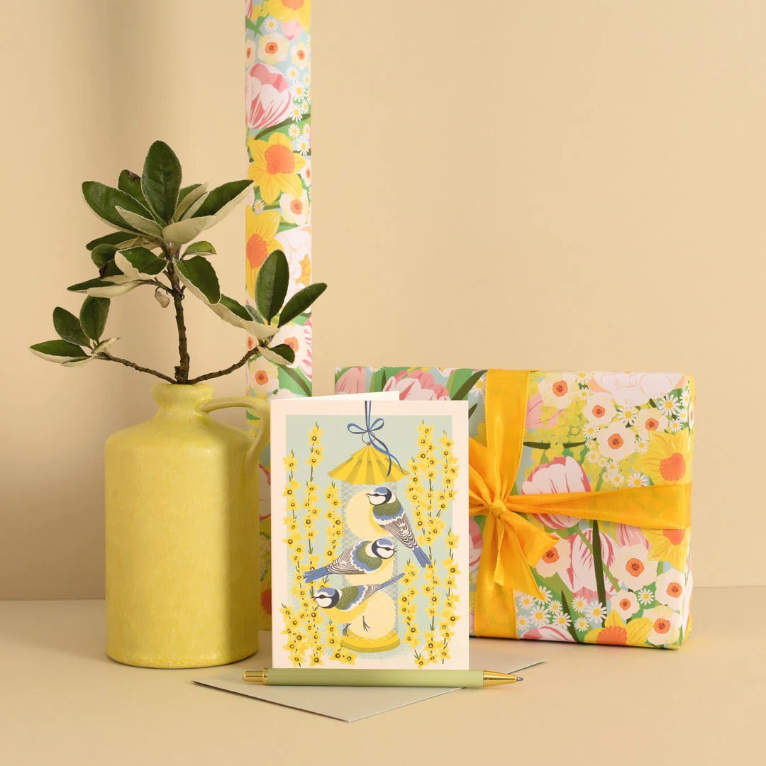 Gift box with floral wrapping paper, card with birds illustration, and yellow vase on a beige background.