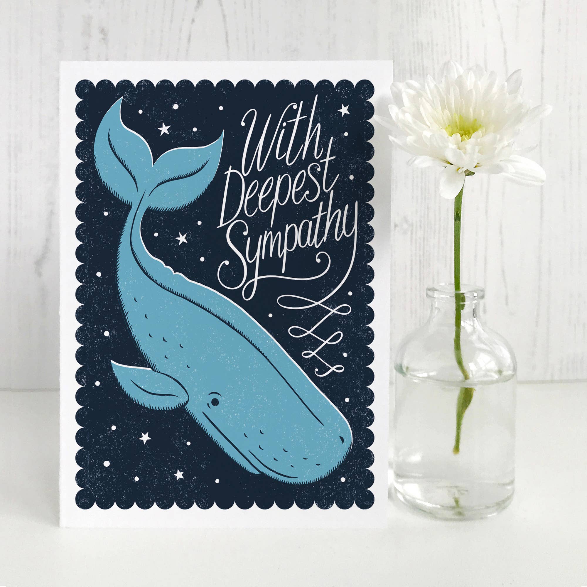 Sympathy card with whale illustration and 'With Deepest Sympathy' text, next to a vase with a white flower on a light wooden surface.