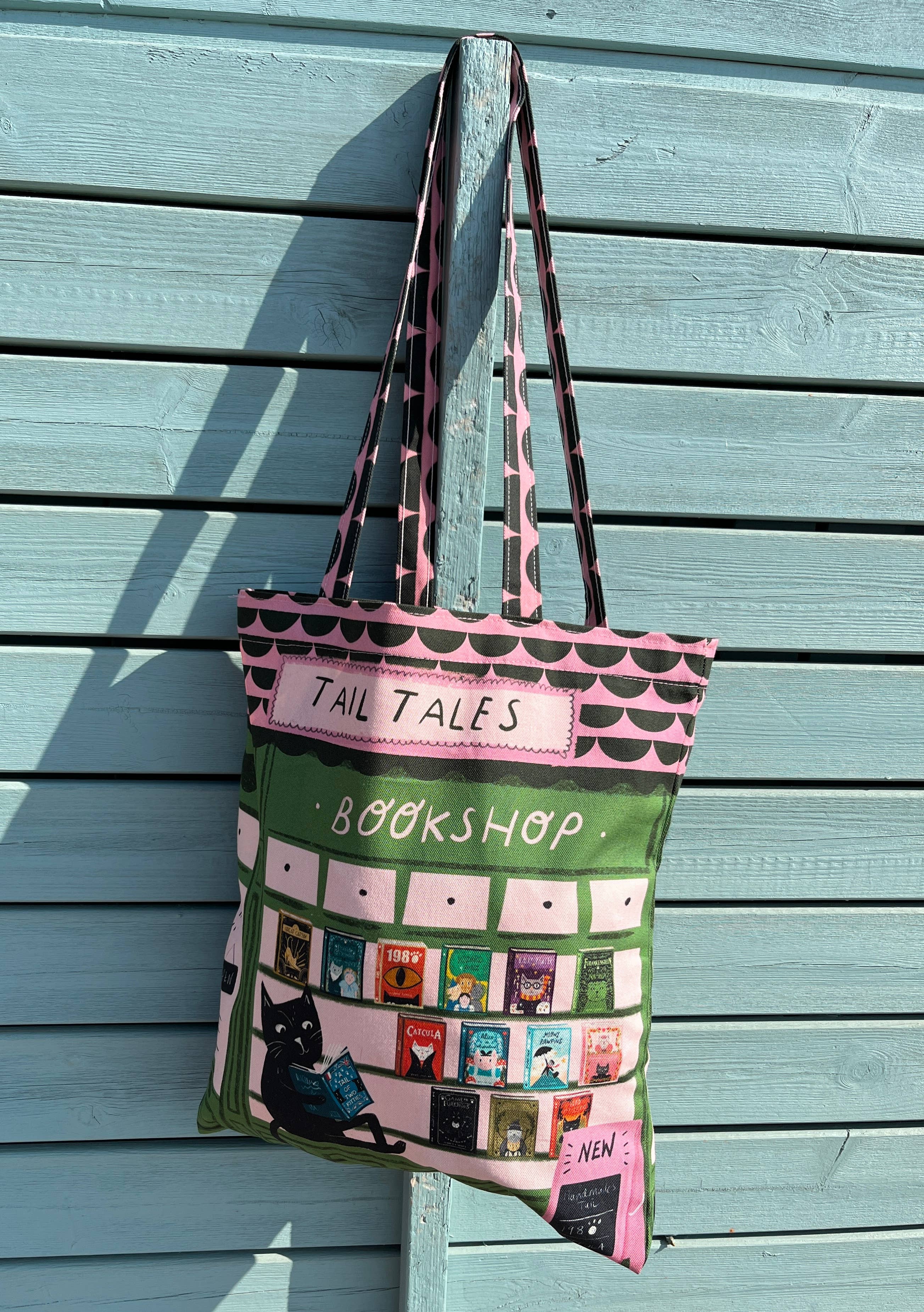 A tote bag with a bookshop theme, featuring a green and pink design with a black cat and book illustrations, hanging from a wooden post against a light blue wooden wall.