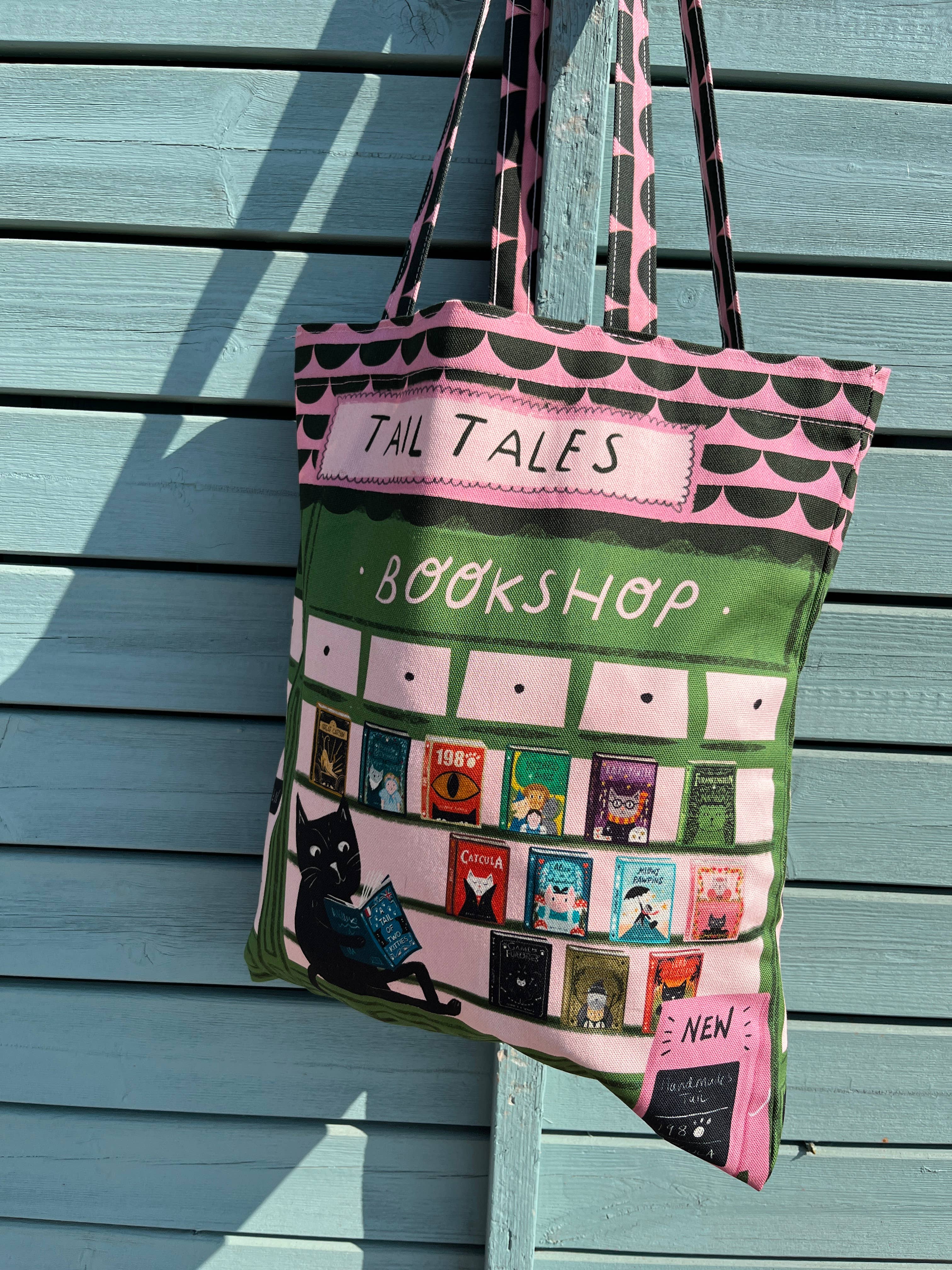 A pink and green fabric tote bag with 'TAIL TALES BOOKSHOP' printed on it, featuring a black cat reading a book and book covers on the front, hanging against a light blue wooden wall.