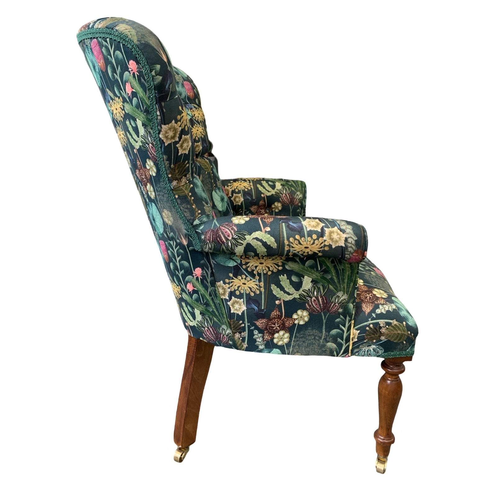 A dark green linen armchair with a turquoise, mustard, pink, and green botanical design, featuring a button-back and mahogany legs with gold castors.