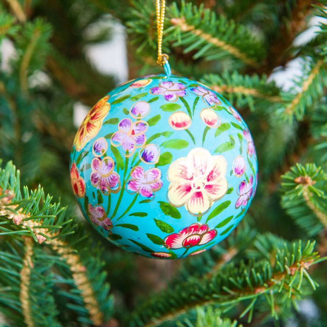 A round papier mache bauble in turquoise with a lilac, yellow and red floral design hanging on a Christmas tree