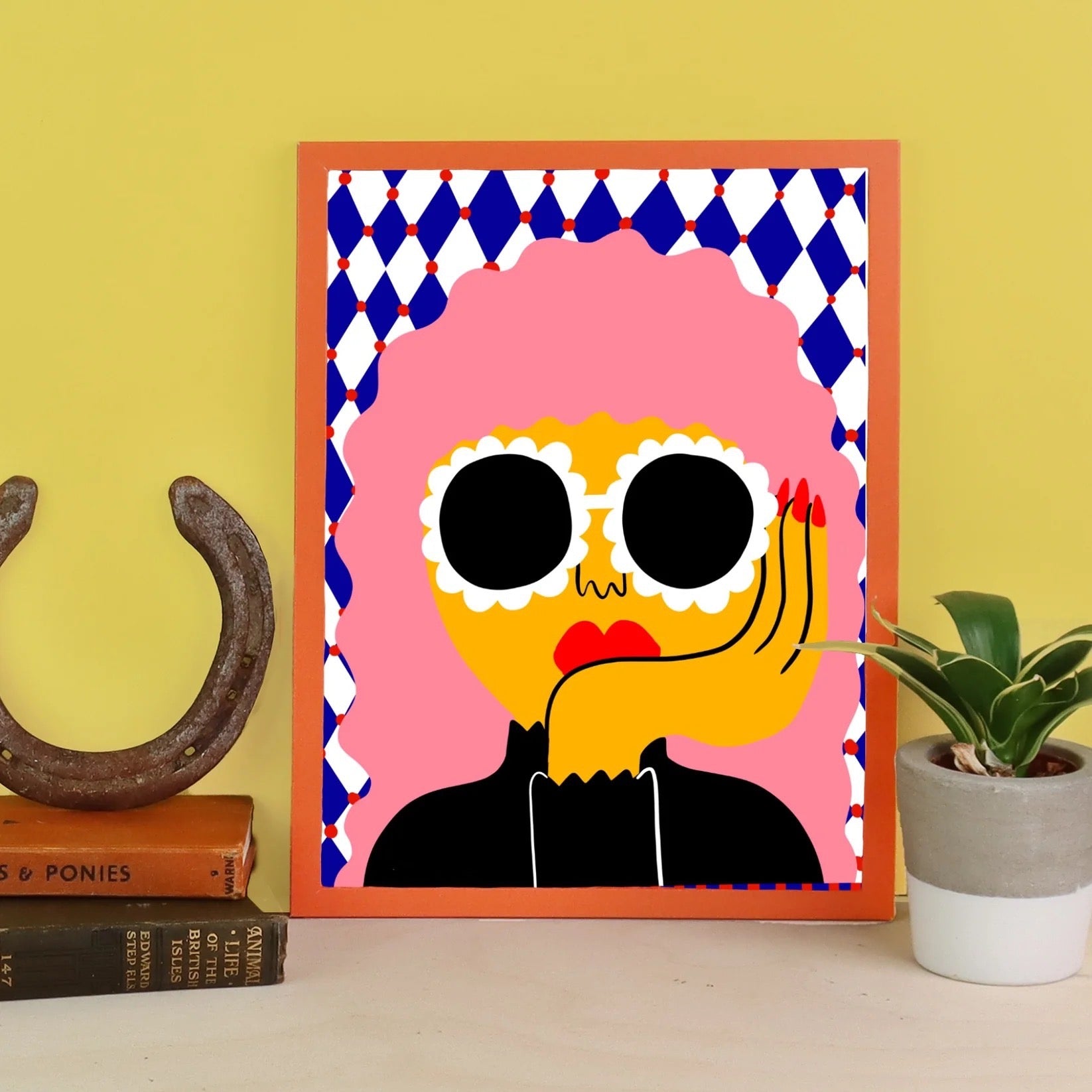 Framed colorful artwork of a stylised face on a yellow wall with books and a plant.