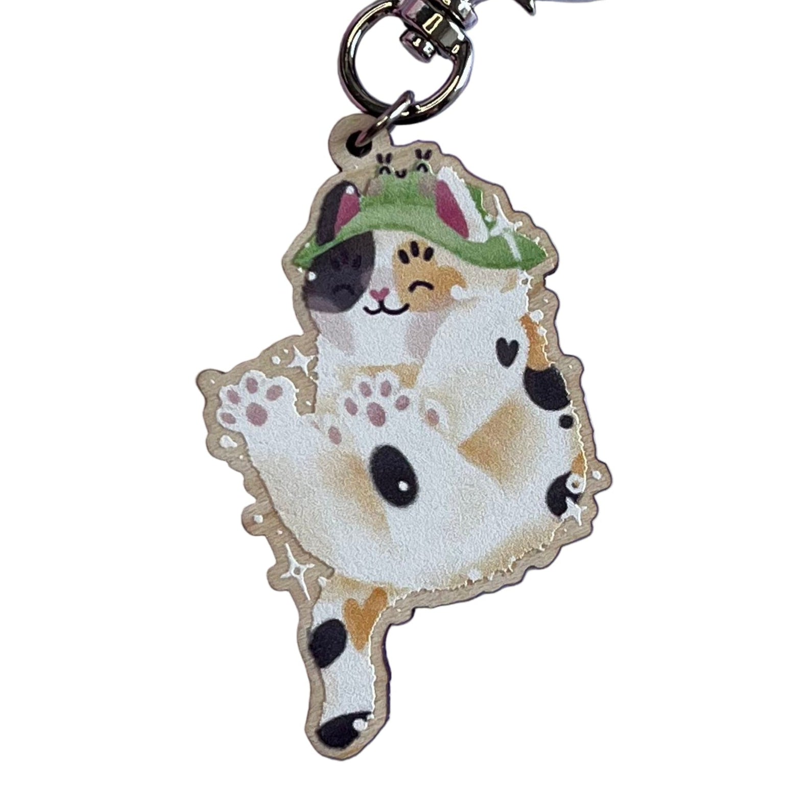 Wooden keyring of a calico cat doing a big stretch, while wearing a frog bucket hat