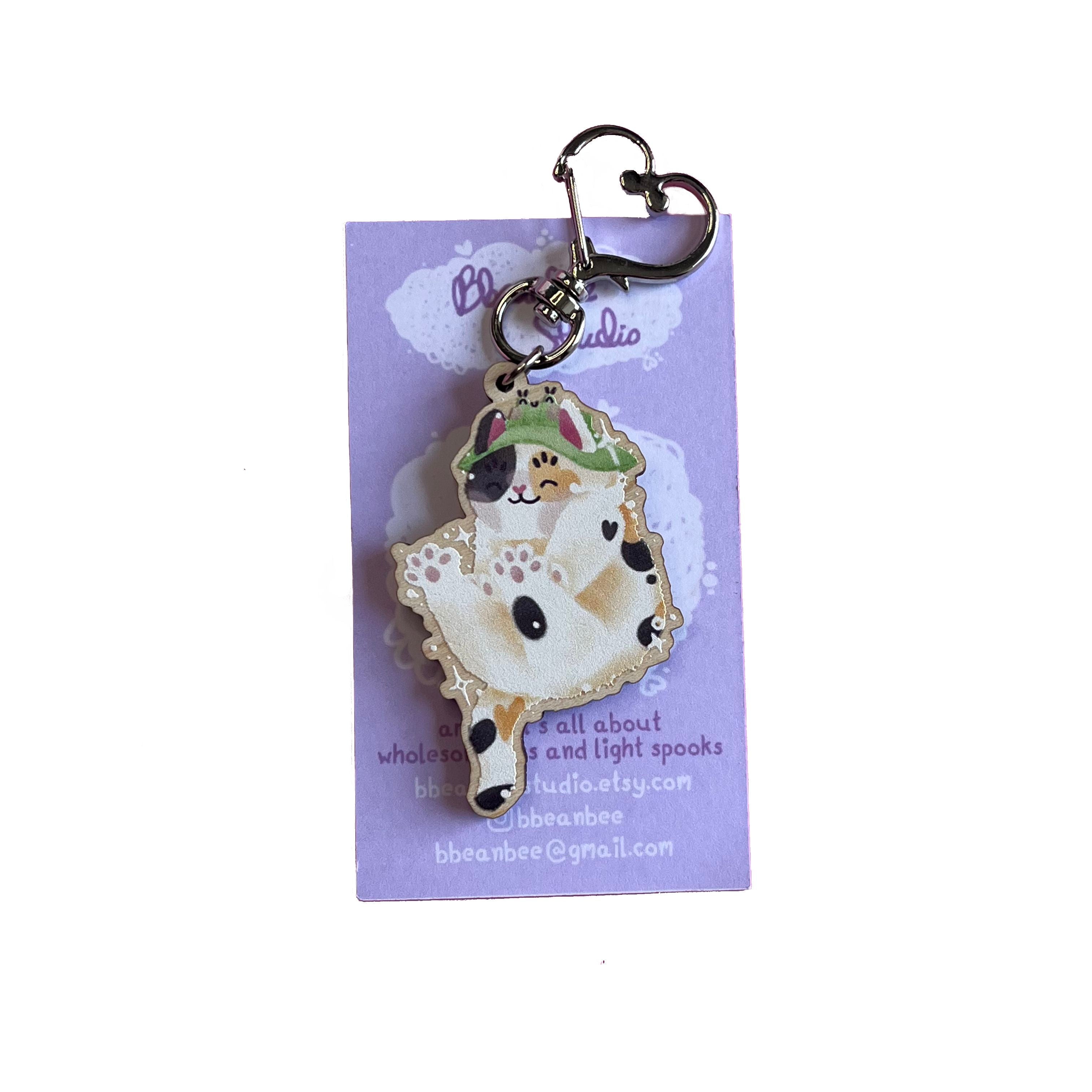 Wooden keyring with silver, heart-shaped lobster clasp. Bucket the Frog Bucket Hat wearing calico cat is doing a big stretch in this keyring design. This keyring is printed onto 4mm thick responsibly sourced European birch plywood.