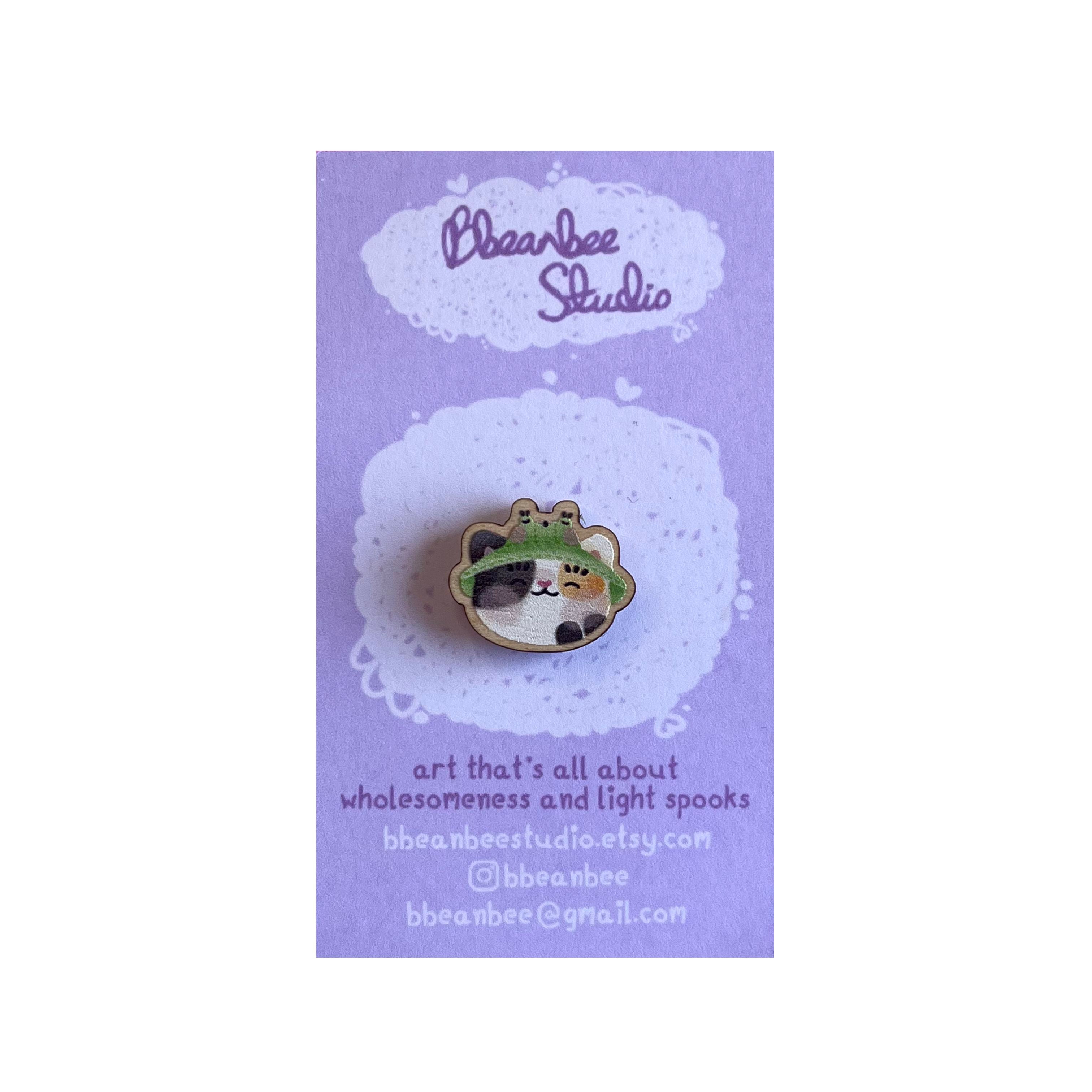 Mini wooden pin badge of a calico cat wearing a frog bucket hat. This badge is printed onto 3mm thick responsibly sourced European birch plywood.