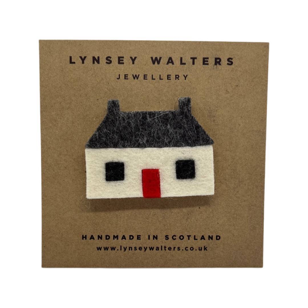 Felt brooch of a traditional Scottish but 'n' ben featuring a white structure with a black roof and windows and a red door.