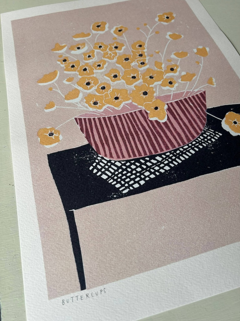 A print featuring a pink vase filled with yellow buttercups, placed on a table with a pink and black striped cloth. The vase and buttercups have a linocut print style.