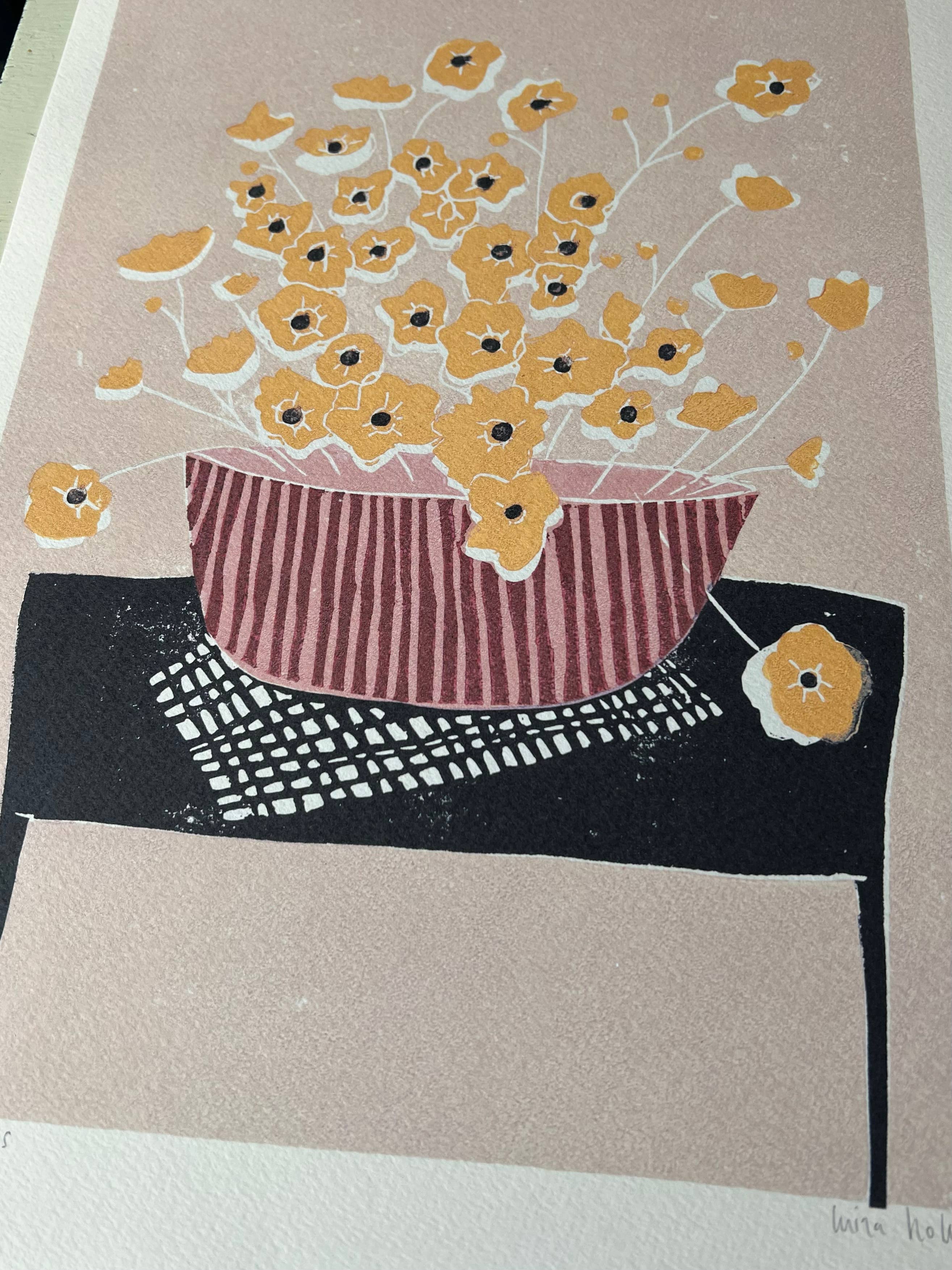 A print featuring a pink vase filled with yellow buttercups, placed on a table with a pink and black striped cloth. The vase and buttercups have a linocut print style.