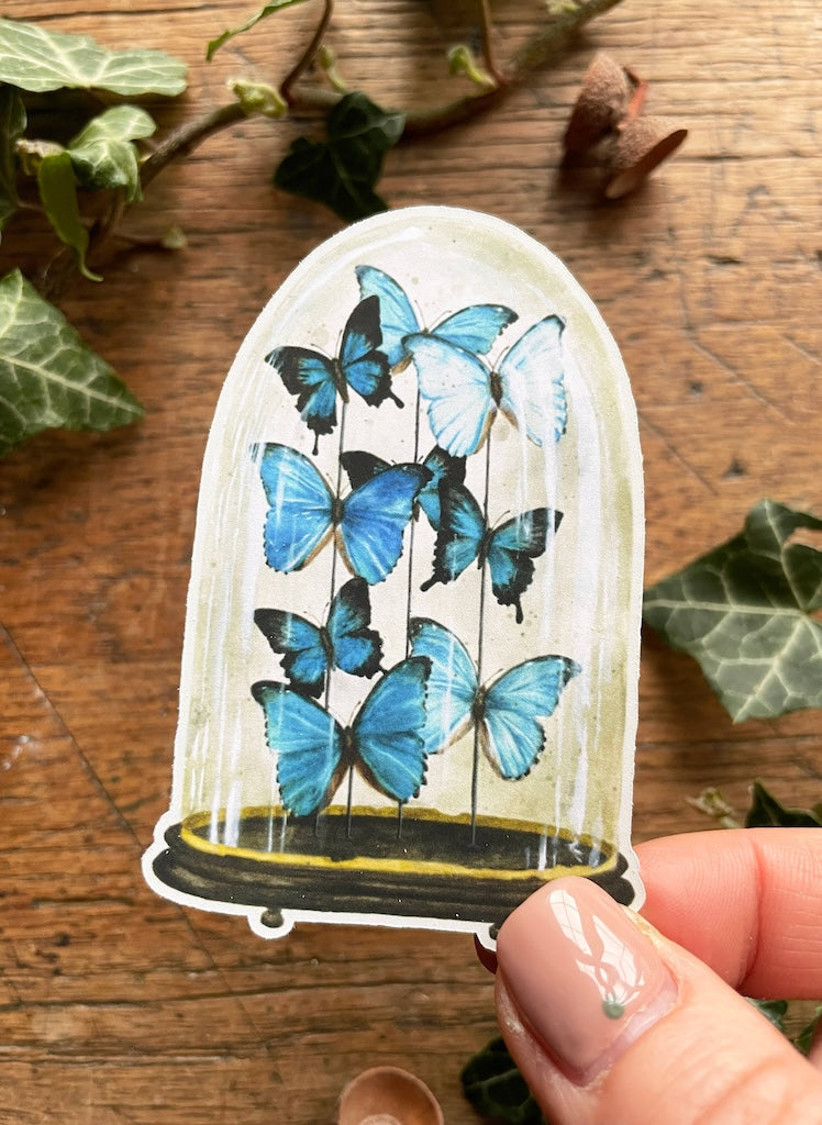Decorative sticker with blue butterflies held by a hand on a wooden surface with ivy leaves.