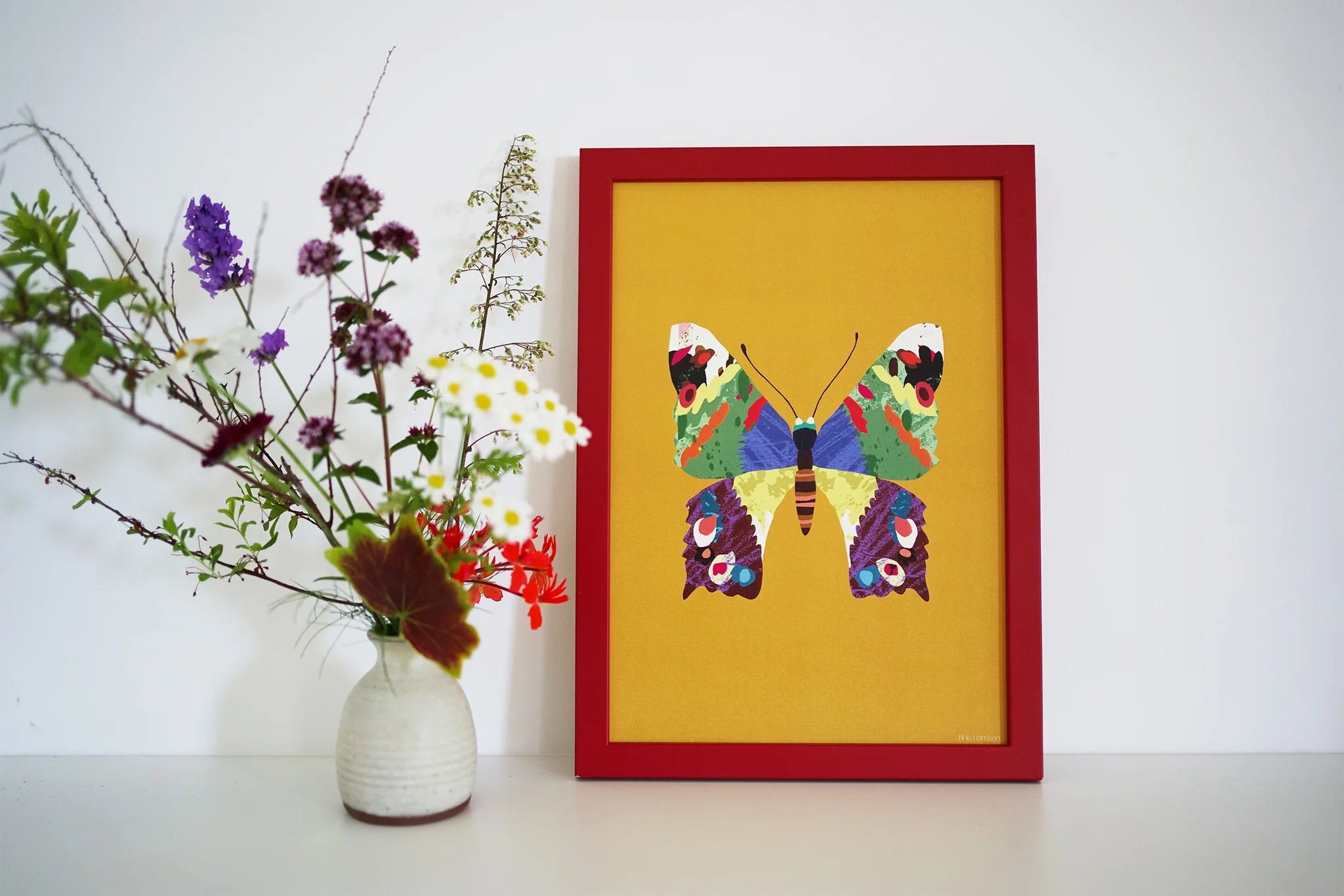 Framed colorful butterfly artwork on a yellow background with a vase of flowers to the left.