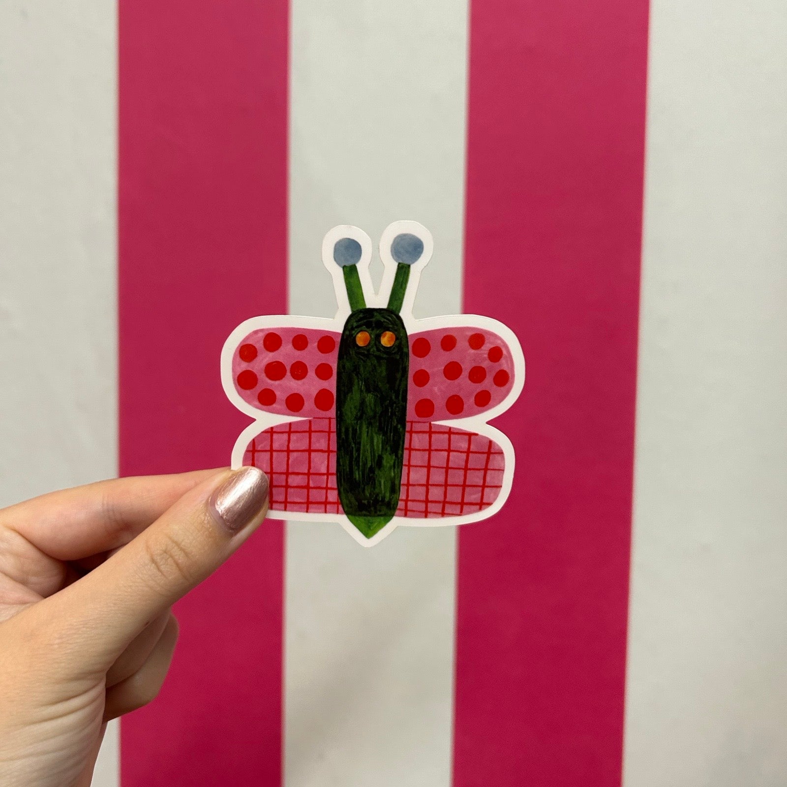 A hand holds a sticker shaped like a stylised butterfly with red polka-dot wings, a red grid patterned lower wing, and a dark green body with blue antennae against a pink and white striped background.